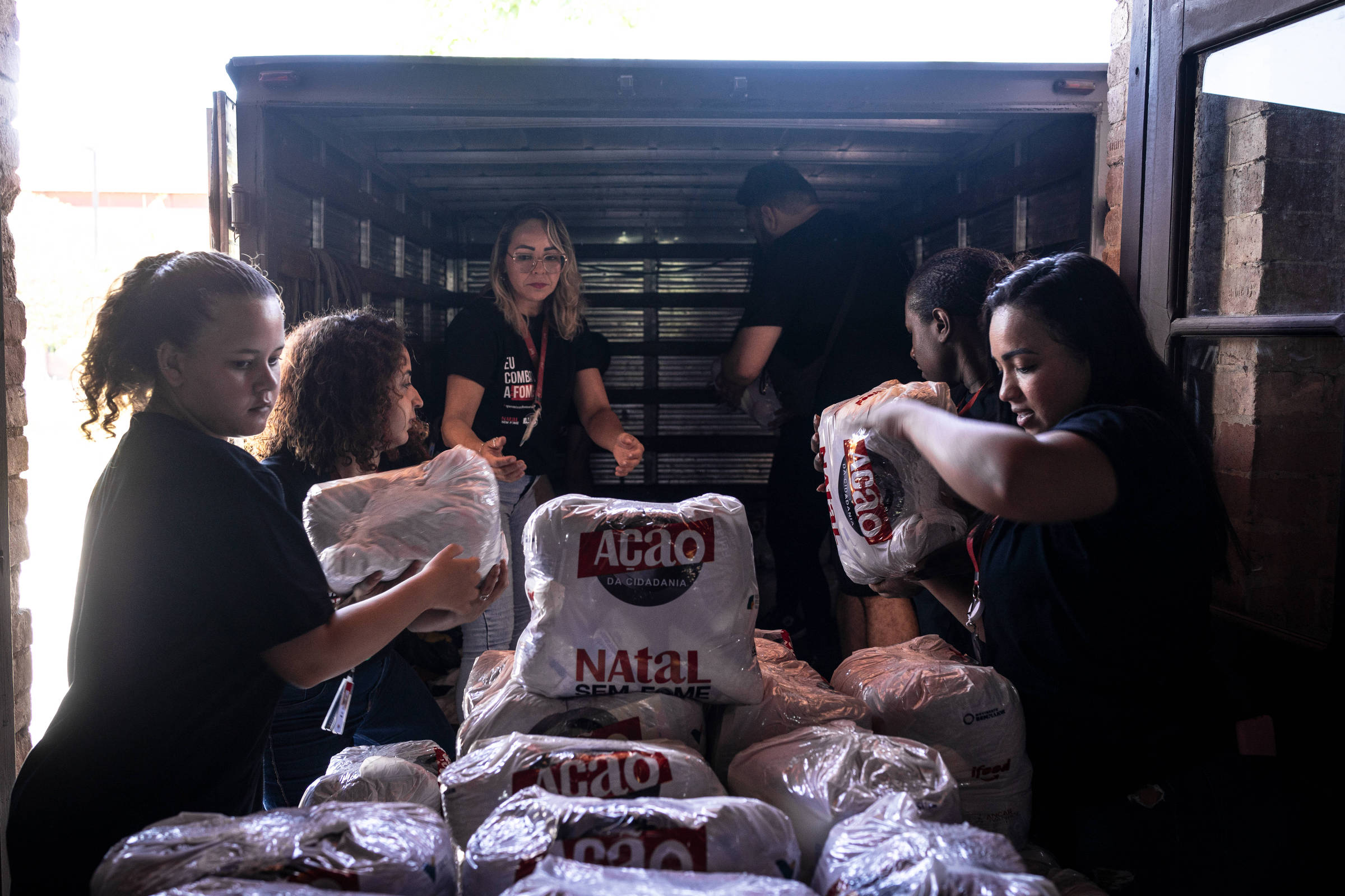 A imagem mostra um grupo de pessoas trabalhando em um caminhão, descarregando pacotes de alimentos. Algumas pessoas estão dentro do caminhão, enquanto outras estão do lado de fora, organizando os pacotes. Todos estão usando camisetas pretas e parecem concentrados na tarefa. Os pacotes têm a marca