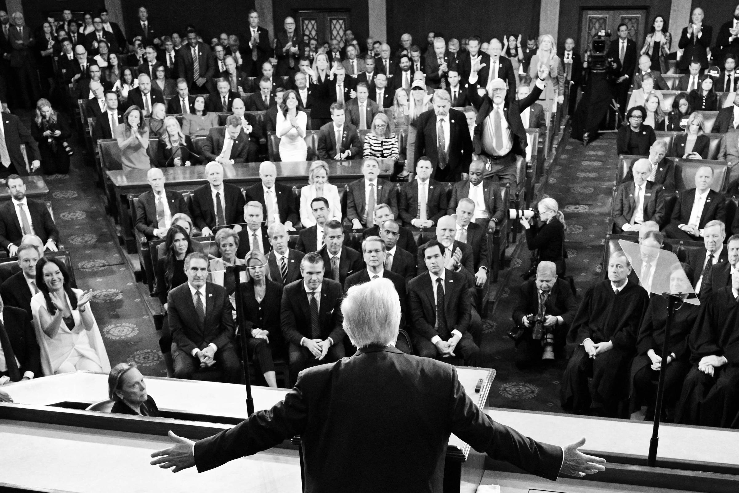 A imagem mostra uma sessão conjunta do Congresso dos Estados Unidos, com um grande número de pessoas presentes. No primeiro plano, um homem de costas, aparentemente falando, com os braços abertos. Atrás dele, há uma plateia composta por membros do Congresso, juízes e outras pessoas, algumas aplaudindo e outras em pé. O ambiente é formal, com bandeiras e símbolos do governo ao fundo.