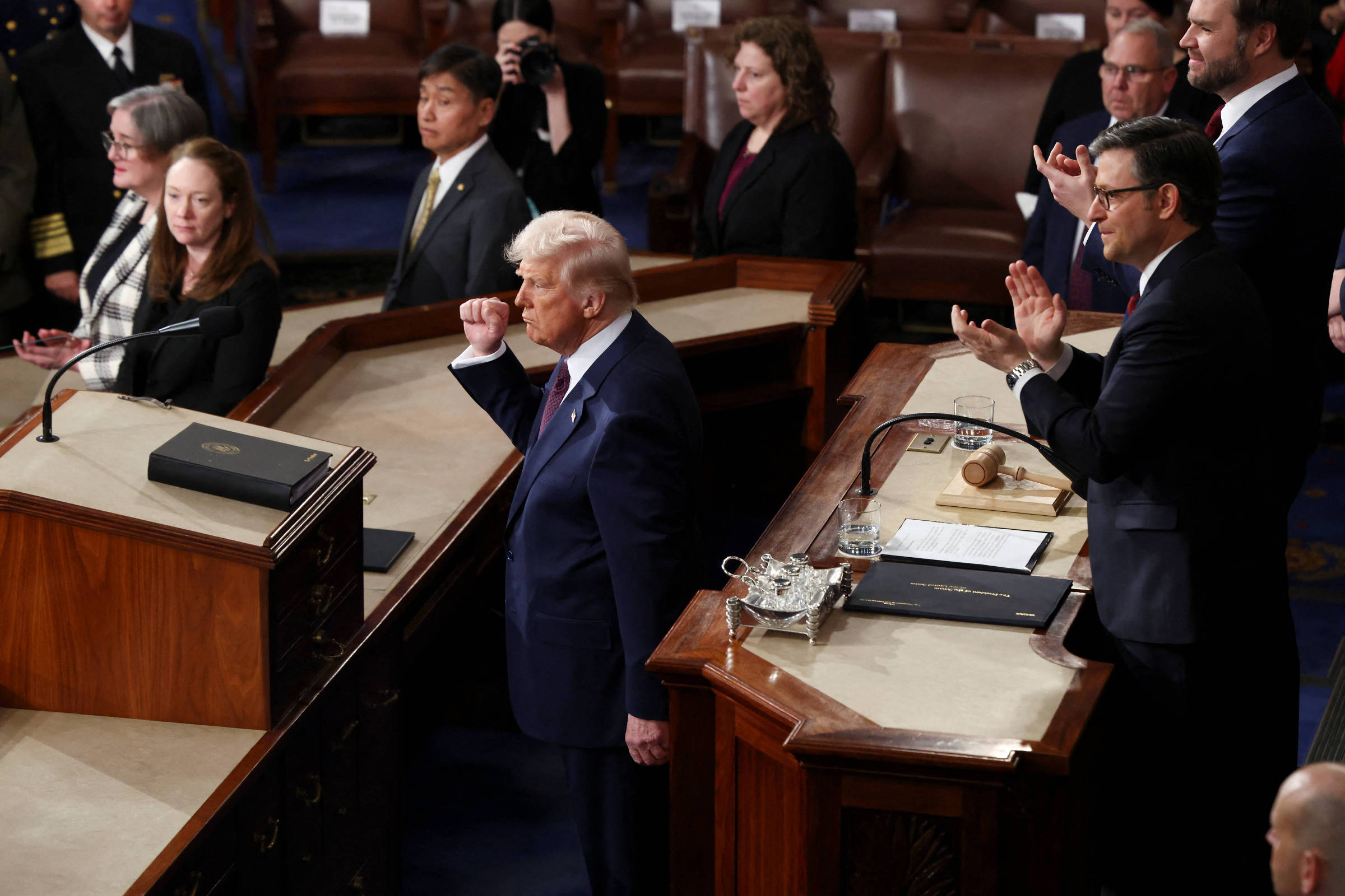 A imagem mostra um momento de um discurso no Congresso dos Estados Unidos. Um homem de cabelo loiro, vestido com um terno escuro, está em pé, fazendo um gesto com a mão. À sua frente, há mesas com documentos e um martelo. Ao fundo, várias pessoas estão assistindo, algumas aplaudindo. O ambiente é formal, com cadeiras de madeira e uma decoração clássica.