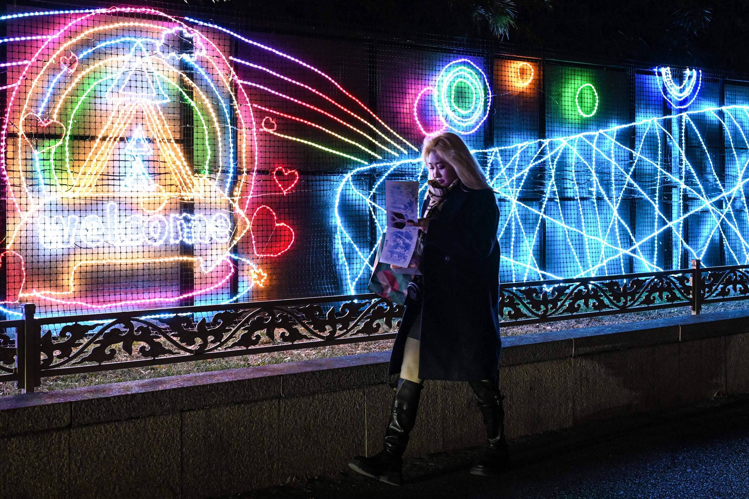 Uma mulher com cabelo longo e liso, vestindo um casaco escuro, caminha ao lado de uma parede iluminada com luzes neon coloridas. As luzes formam padrões vibrantes, incluindo um símbolo de coração e a palavra