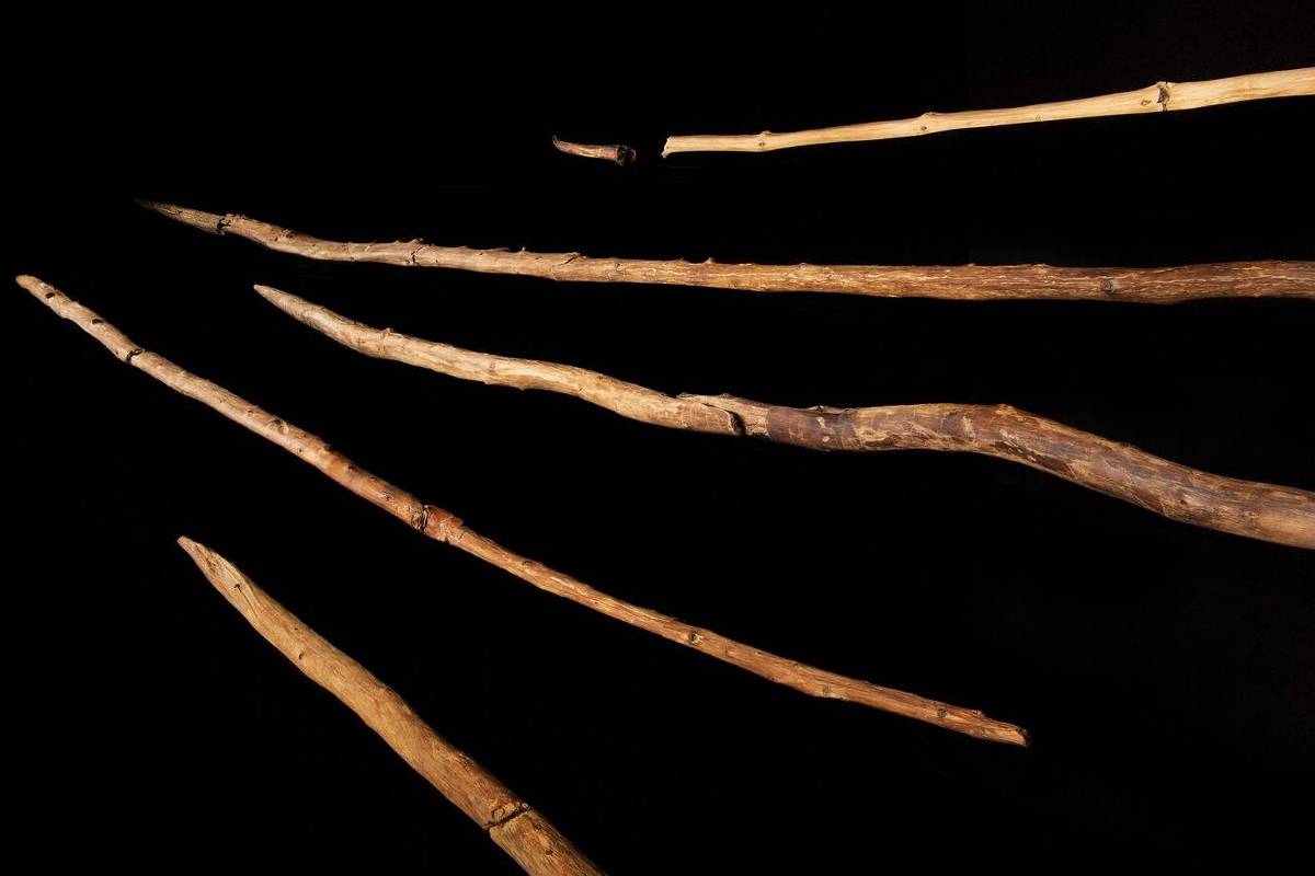 A imagem apresenta várias varas de madeira dispostas em uma superfície escura. As varas têm diferentes comprimentos e formas, com algumas apresentando extremidades afiadas. O fundo é preto, destacando as varas em primeiro plano.