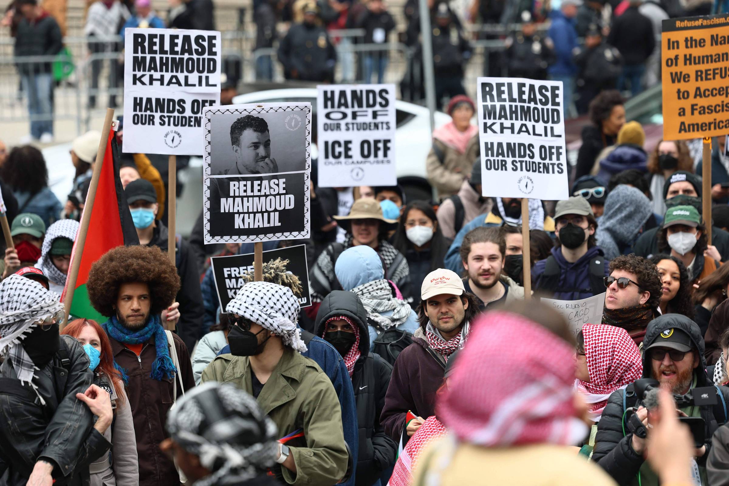 A imagem mostra uma manifestação com várias pessoas segurando cartazes. Os cartazes têm os dizeres