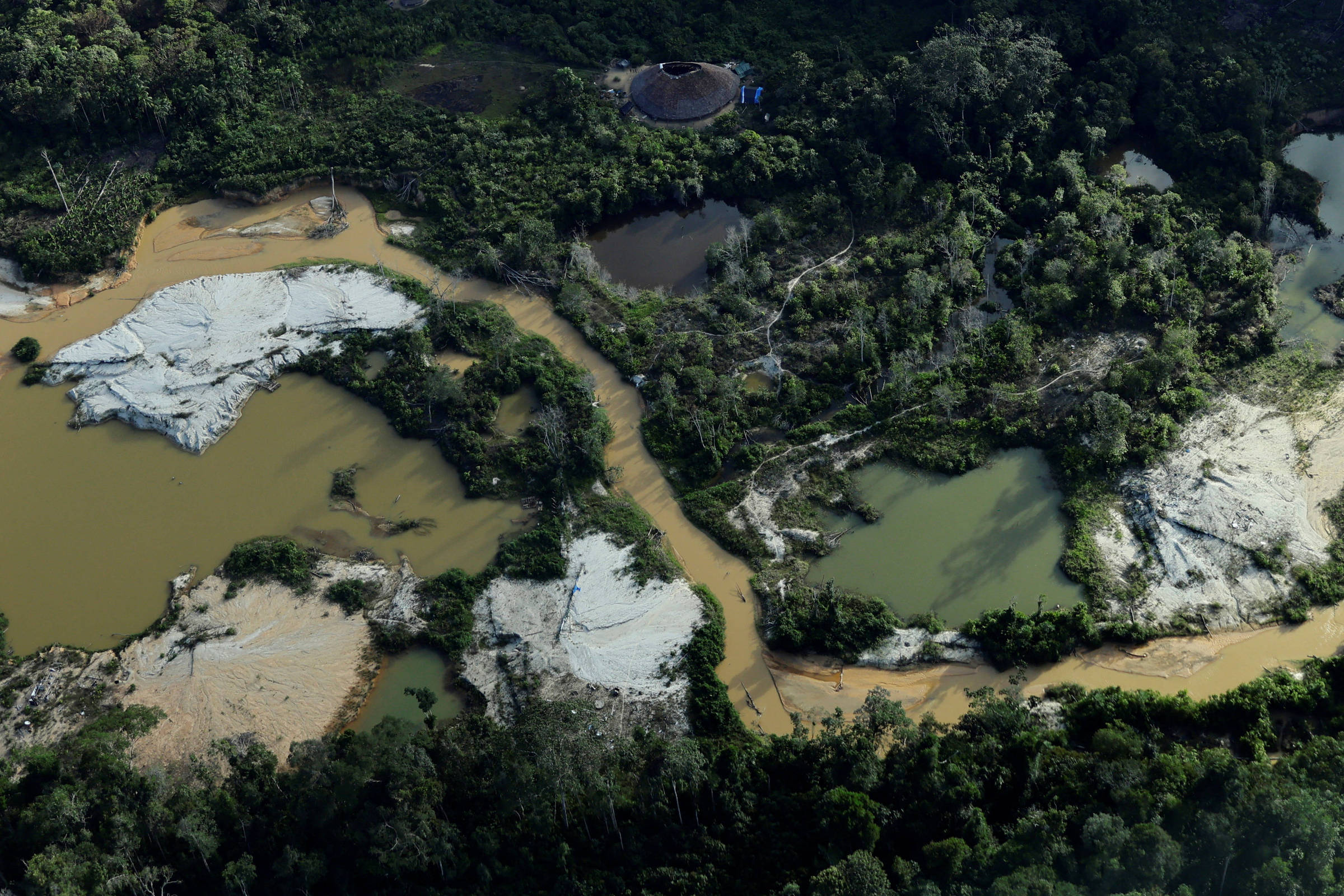 A imagem aérea mostra uma área de floresta amazônica com várias lagoas e rios. As águas estão em diferentes tonalidades, variando entre marrom e verde, indicando poluição e desmatamento. Há também áreas de solo exposto, onde a vegetação foi removida, e uma estrutura visível ao fundo, possivelmente uma instalação industrial.