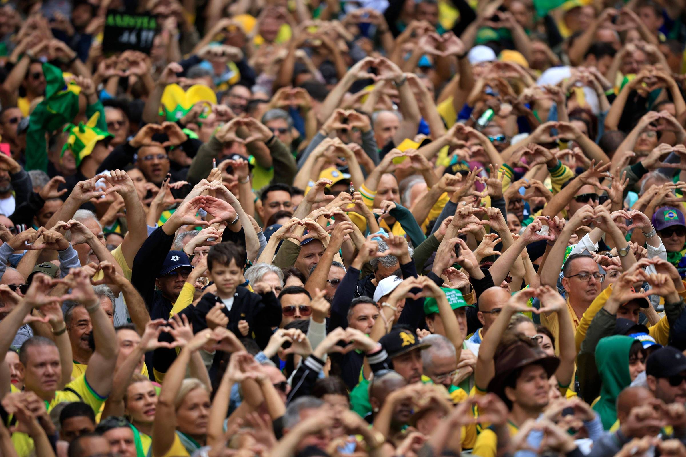 Uma grande multidão de pessoas está reunida, todas fazendo gestos de coração com as mãos. A maioria das pessoas está vestida com roupas nas cores verde e amarelo, representando o Brasil. O ambiente é festivo, com algumas pessoas segurando celulares e outras com bandeiras. O público parece animado e engajado.