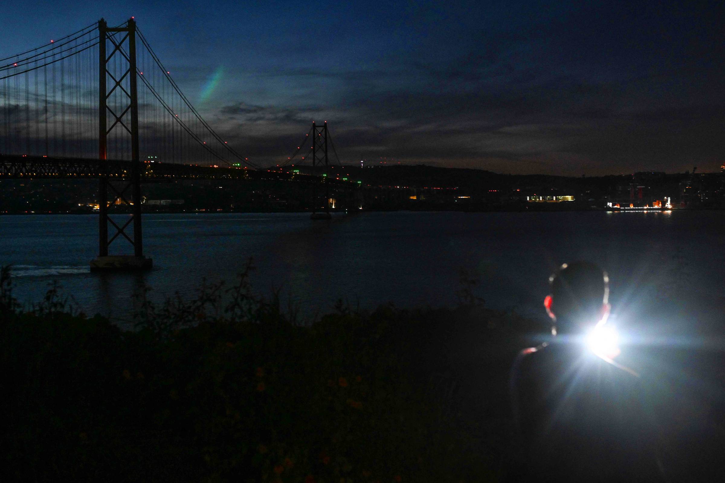 A imagem mostra uma pessoa de costas, segurando uma lanterna que emite uma luz forte. Ao fundo, é visível uma ponte iluminada sobre um rio, com a cidade ao longe. O céu está escuro, com algumas nuvens e uma leve iluminação na parte superior, sugerindo a presença de estrelas ou uma aurora. A cena transmite uma atmosfera tranquila e noturna.