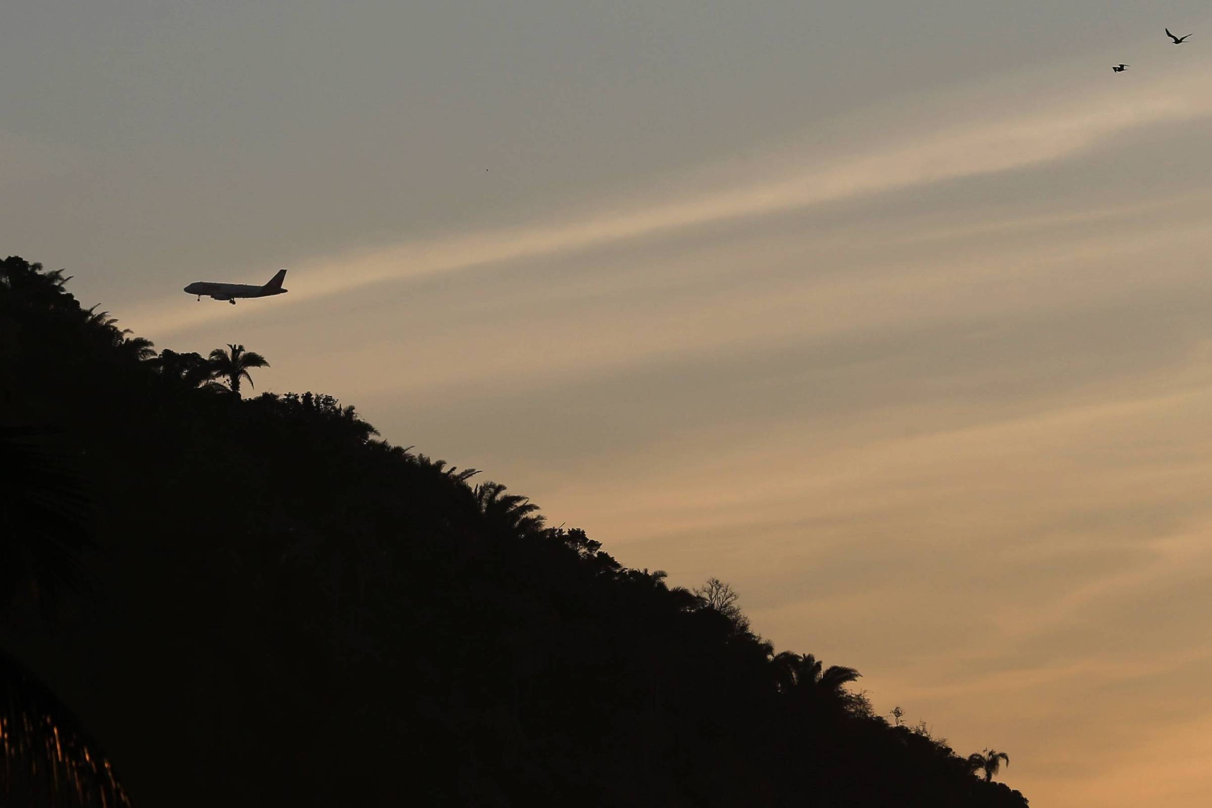 A imagem mostra um avião voando baixo sobre uma montanha, com silhuetas de palmeiras à esquerda. O céu apresenta um tom alaranjado e azul, típico do pôr do sol, e há algumas aves voando no canto superior direito da imagem.