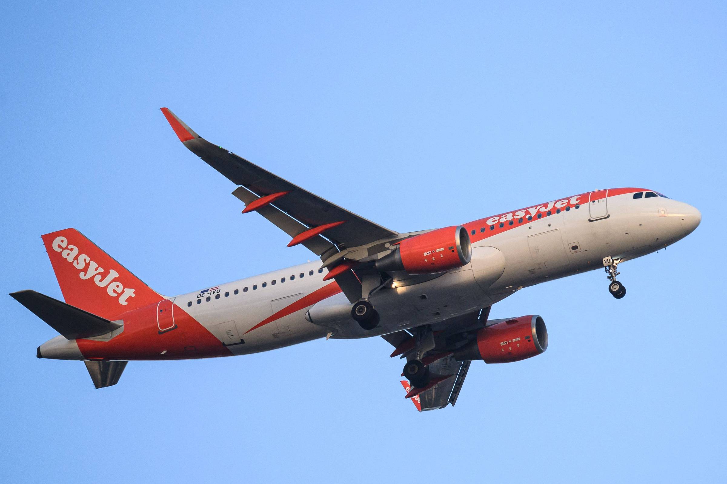 Um avião da companhia aérea EasyJet está voando no céu azul. O avião é de fuselagem branca com detalhes em laranja, incluindo o logotipo da EasyJet na lateral. As asas são longas e possuem flaps visíveis, enquanto os motores estão localizados sob as asas.