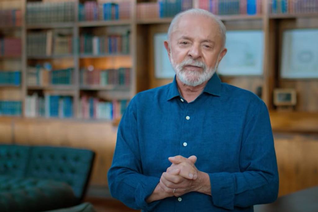Um homem idoso com cabelo grisalho e barba, vestindo uma camisa azul, está em pé com as mãos unidas em frente ao corpo. Ao fundo, há uma estante de livros com várias prateleiras cheias de livros e documentos. O ambiente parece ser uma biblioteca ou um escritório bem iluminado, com um sofá verde em primeiro plano.