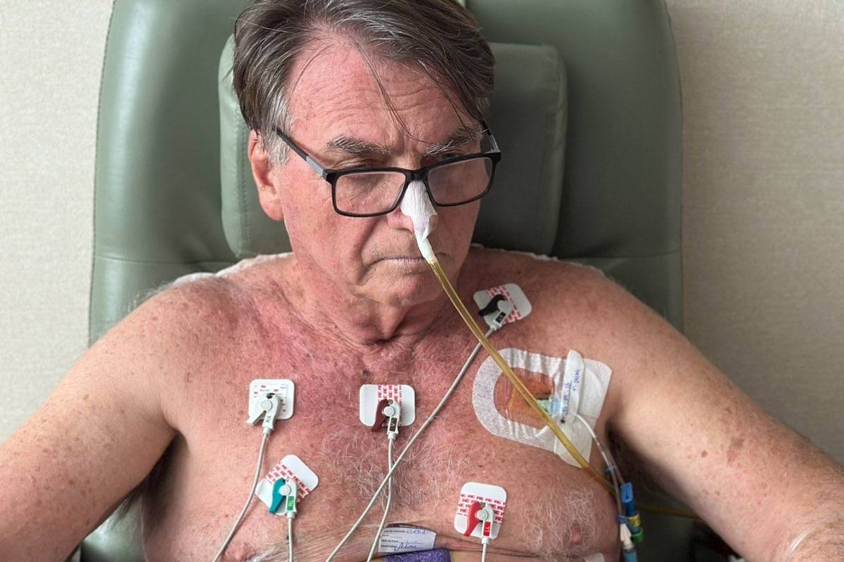 Um homem idoso está sentado em uma cadeira de hospital, com o tronco nu e vários eletrodos adesivos colados ao peito e ao abdômen. Ele usa óculos e tem um tubo de oxigênio inserido no nariz. O homem está segurando uma folha de papel e parece estar concentrado na leitura. O ambiente é de um hospital, com paredes claras e uma cadeira de tratamento.