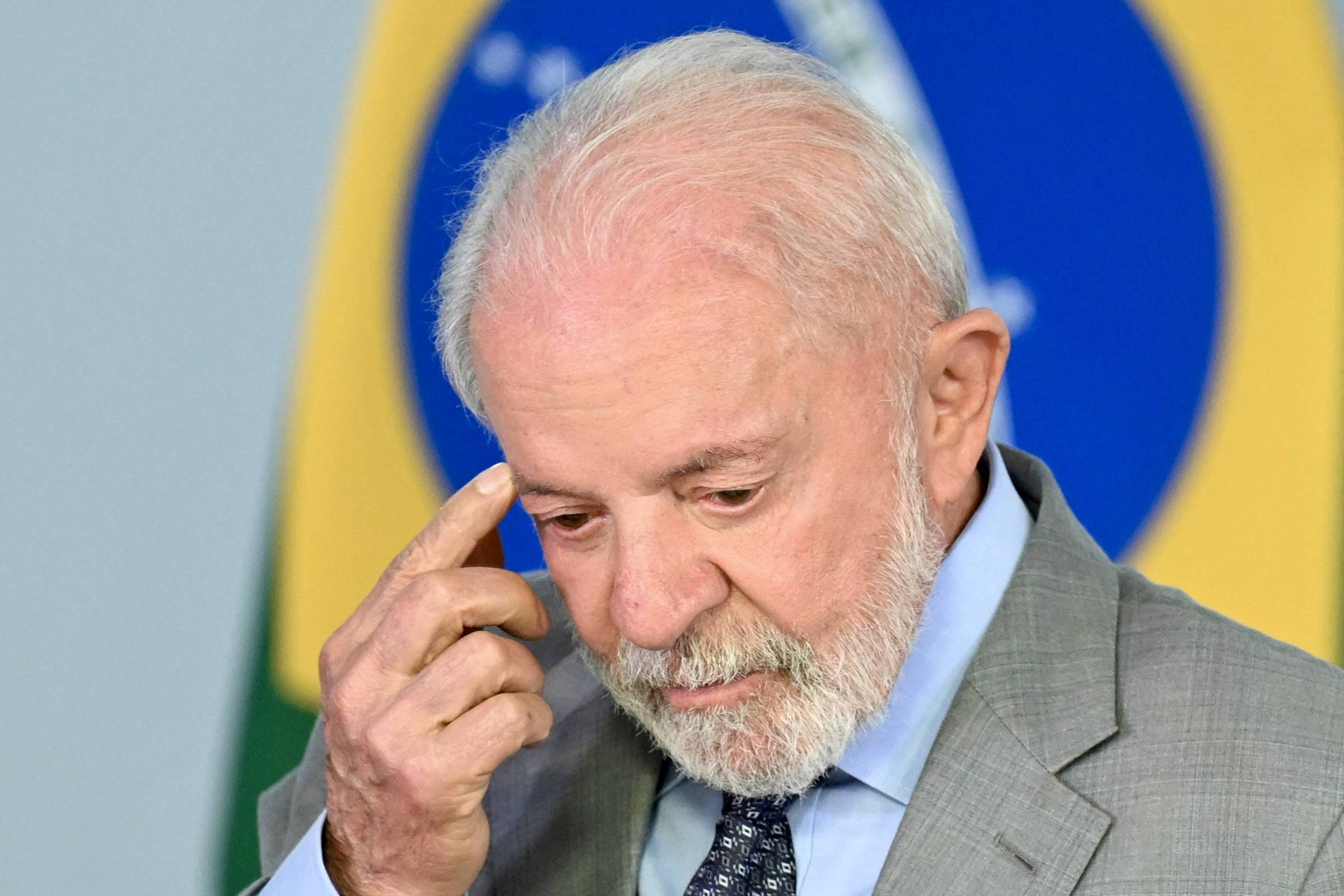 Um homem de cabelo grisalho e barba, vestindo um terno cinza e uma gravata azul, está com a mão na cabeça, aparentando estar pensativo ou reflexivo. Ao fundo, há uma bandeira do Brasil e um símbolo que representa o país.