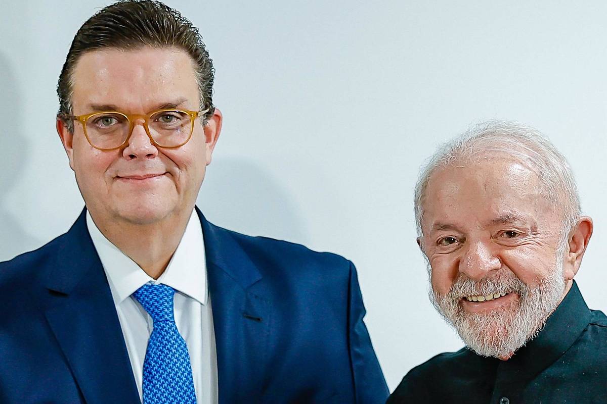 Na imagem, duas pessoas estão posando para a foto, segurando um documento. À esquerda, um homem de terno azul e gravata, com óculos, está sorrindo. À direita, um homem mais velho com barba branca, vestindo uma camisa preta, também sorri. O documento que eles seguram tem uma borda colorida, com as cores verde, amarelo e vermelho, e parece ser um contrato ou um acordo oficial. O fundo é neutro e claro.