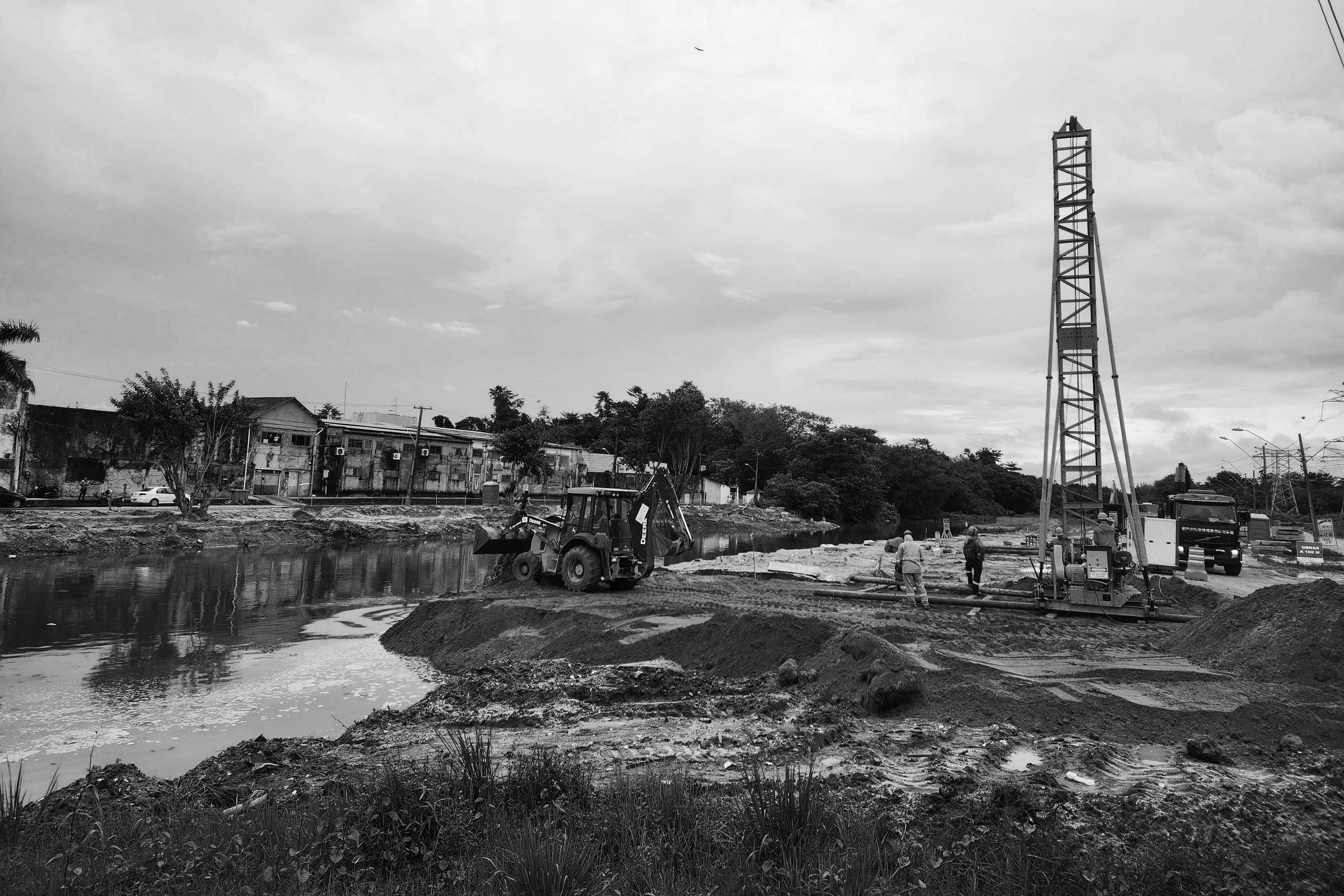 A imagem mostra uma área de construção com um terreno de terra vermelha e uma poça de água. Há uma escavadeira amarela e um guindaste amarelo em operação. Algumas pessoas estão trabalhando na área, e ao fundo, há edifícios e vegetação. O céu está nublado, indicando possibilidade de chuva.