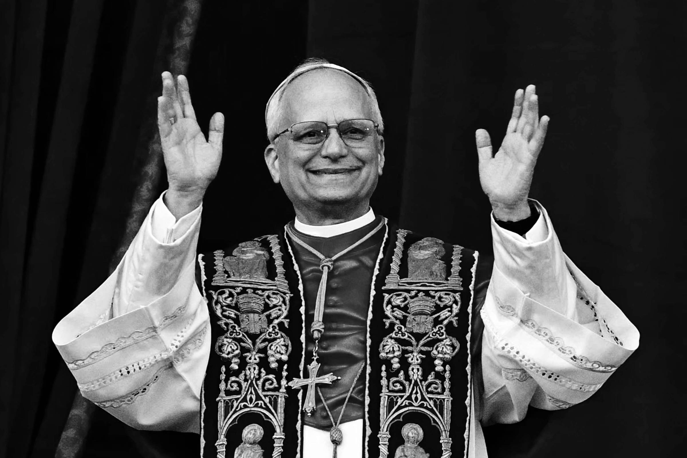 Um homem em vestes religiosas, com uma túnica branca e um manto vermelho decorado com símbolos religiosos. Ele está sorrindo e levantando as mãos em um gesto de saudação. Ao fundo, há um pano vermelho.