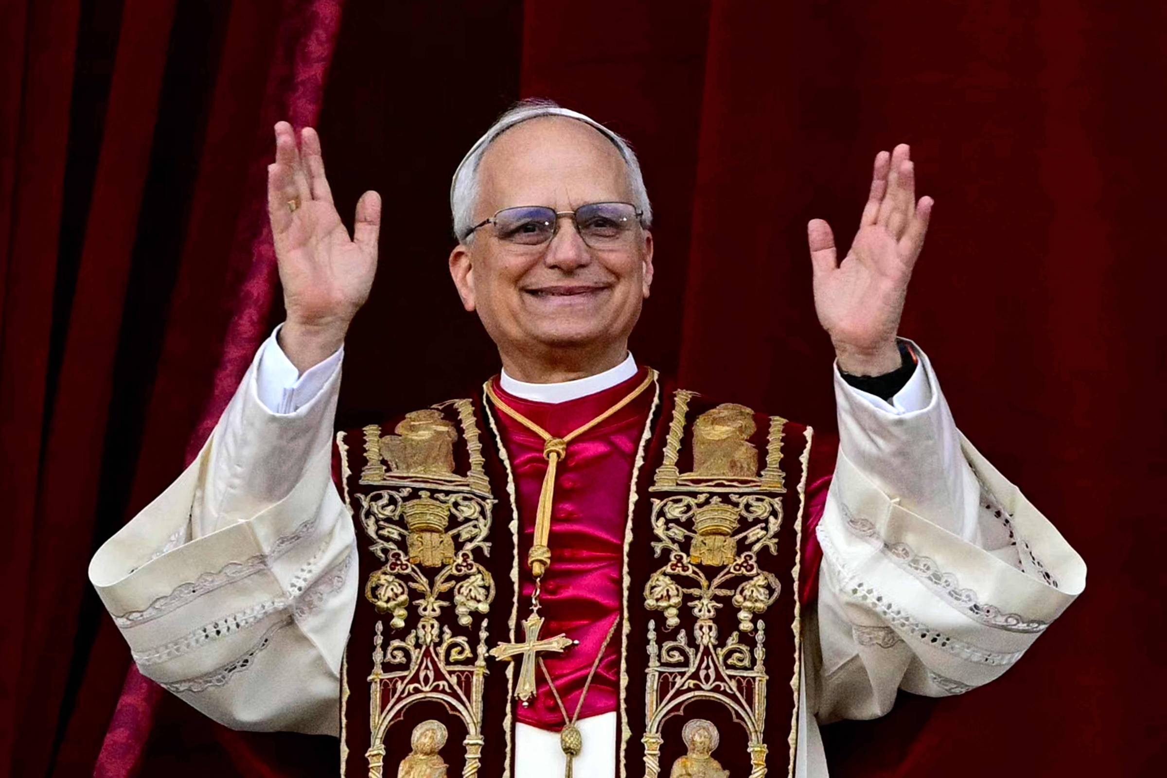Um homem em vestes religiosas, com uma túnica branca e um manto vermelho decorado com símbolos religiosos. Ele está sorrindo e levantando as mãos em um gesto de saudação. Ao fundo, há um pano vermelho.