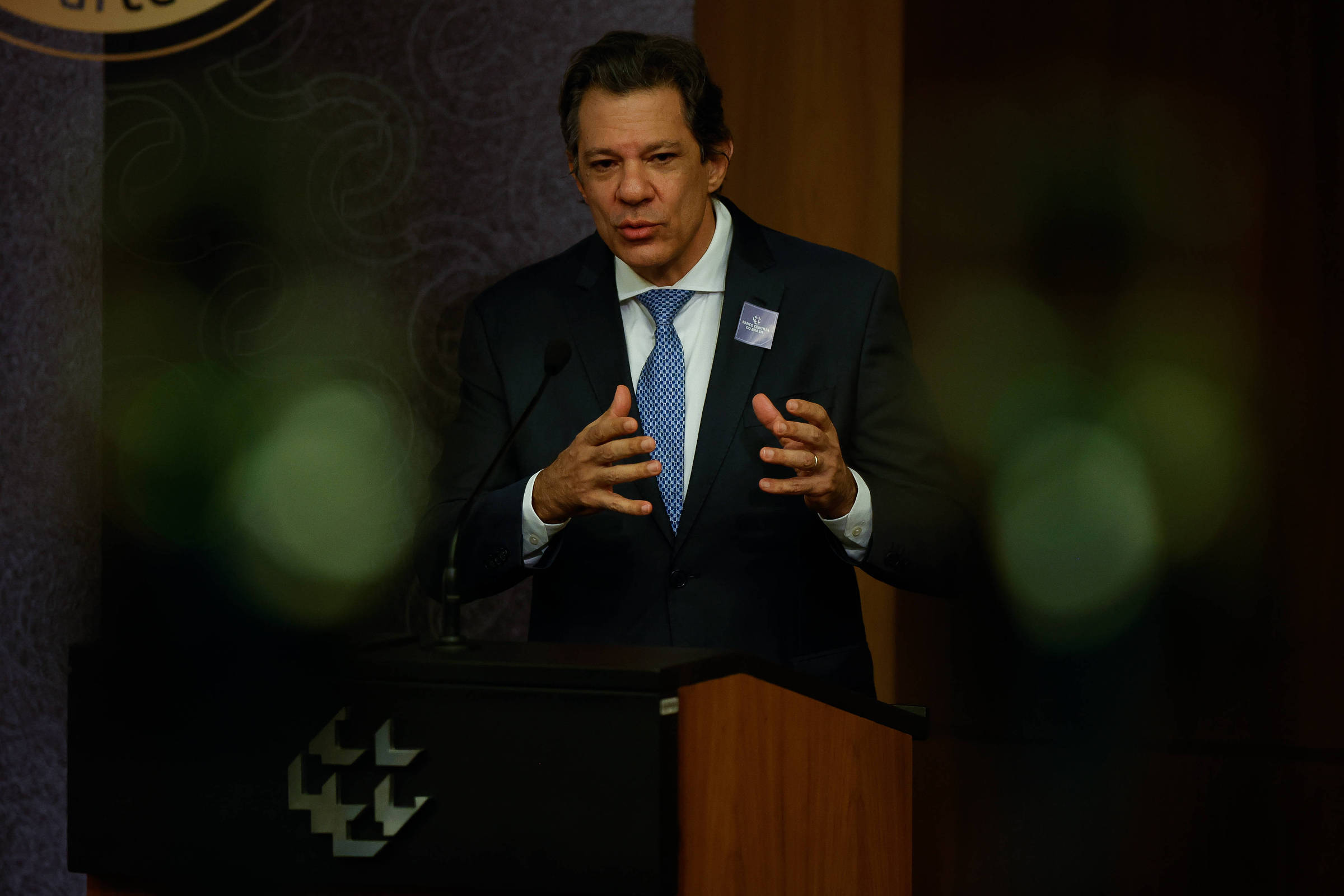 Imagem mostra Fernando Haddad, um homem em um terno escuro e que está falando em um microfone em um evento. Ele gesticula com as mãos enquanto se expressa. Ao fundo, há uma mesa com um logotipo visível e uma decoração em tons de marrom e dourado. A imagem tem um foco suave em algumas áreas, criando um efeito de desfoque em primeiro plano.