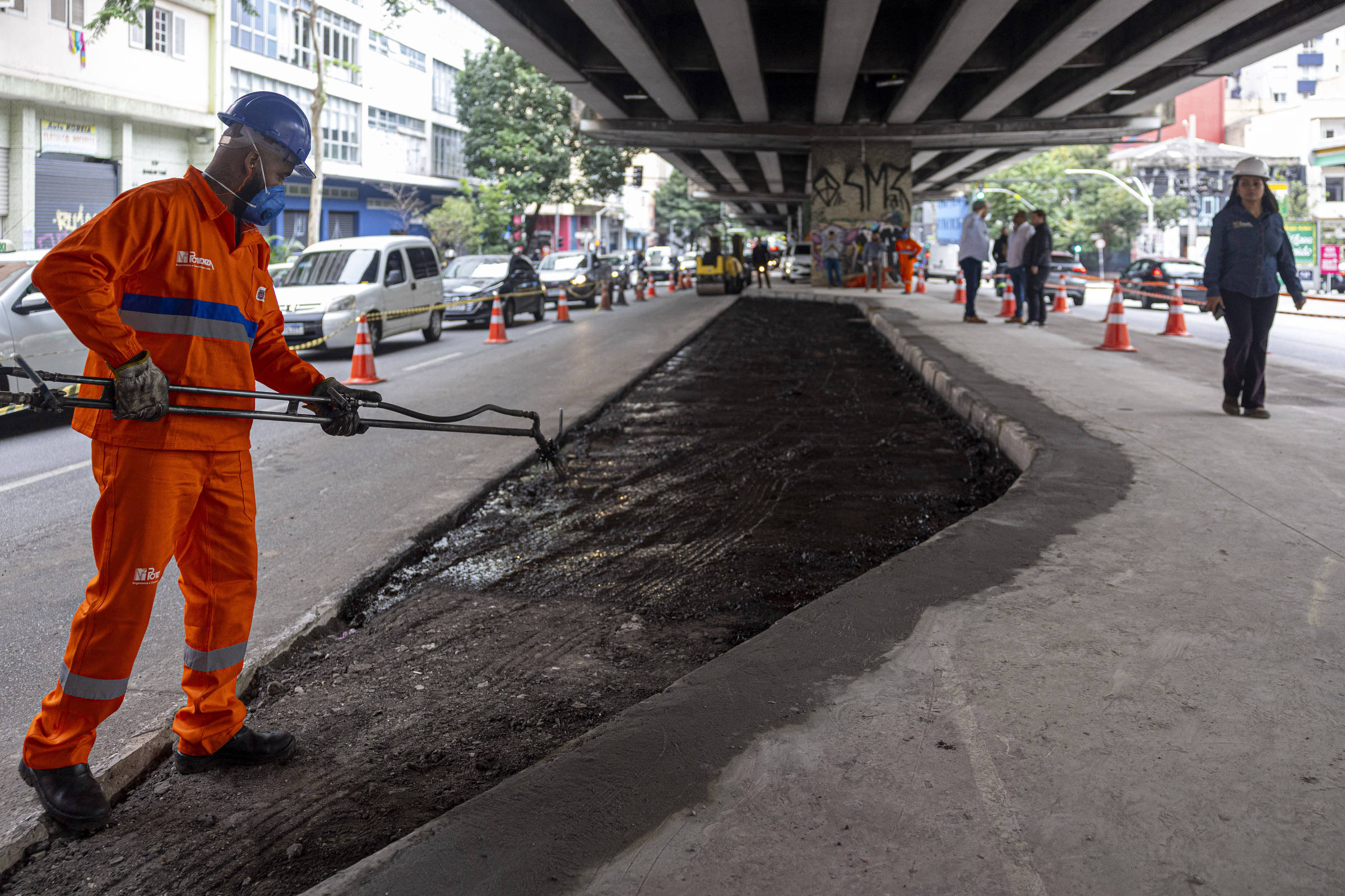 A imagem mostra um trabalhador vestido com um uniforme laranja e capacete, utilizando uma ferramenta para realizar reparos em uma pista sob um viaduto. O local está cercado por cones de sinalização e há outros trabalhadores ao fundo. Vários veículos estão estacionados ao lado da obra, e o ambiente é urbano, com prédios visíveis ao fundo.