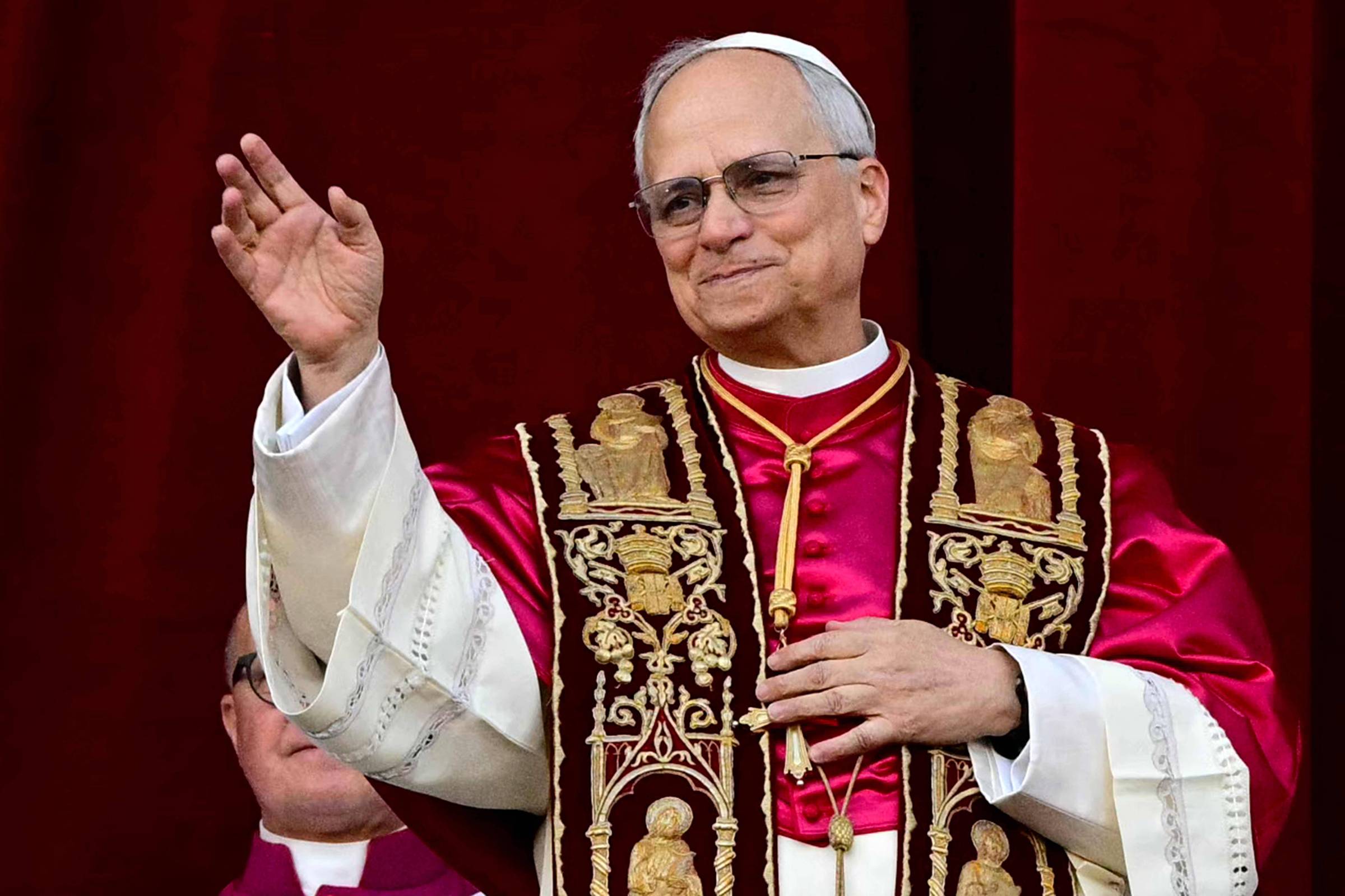 Um homem com óculos e cabelo grisalho, vestido com vestes religiosas vermelhas e brancas, acena com a mão direita. Ele está em um ambiente formal, possivelmente em uma cerimônia religiosa, com um fundo vermelho. Ao fundo, é possível ver outra pessoa, parcialmente visível, também vestida de maneira formal.