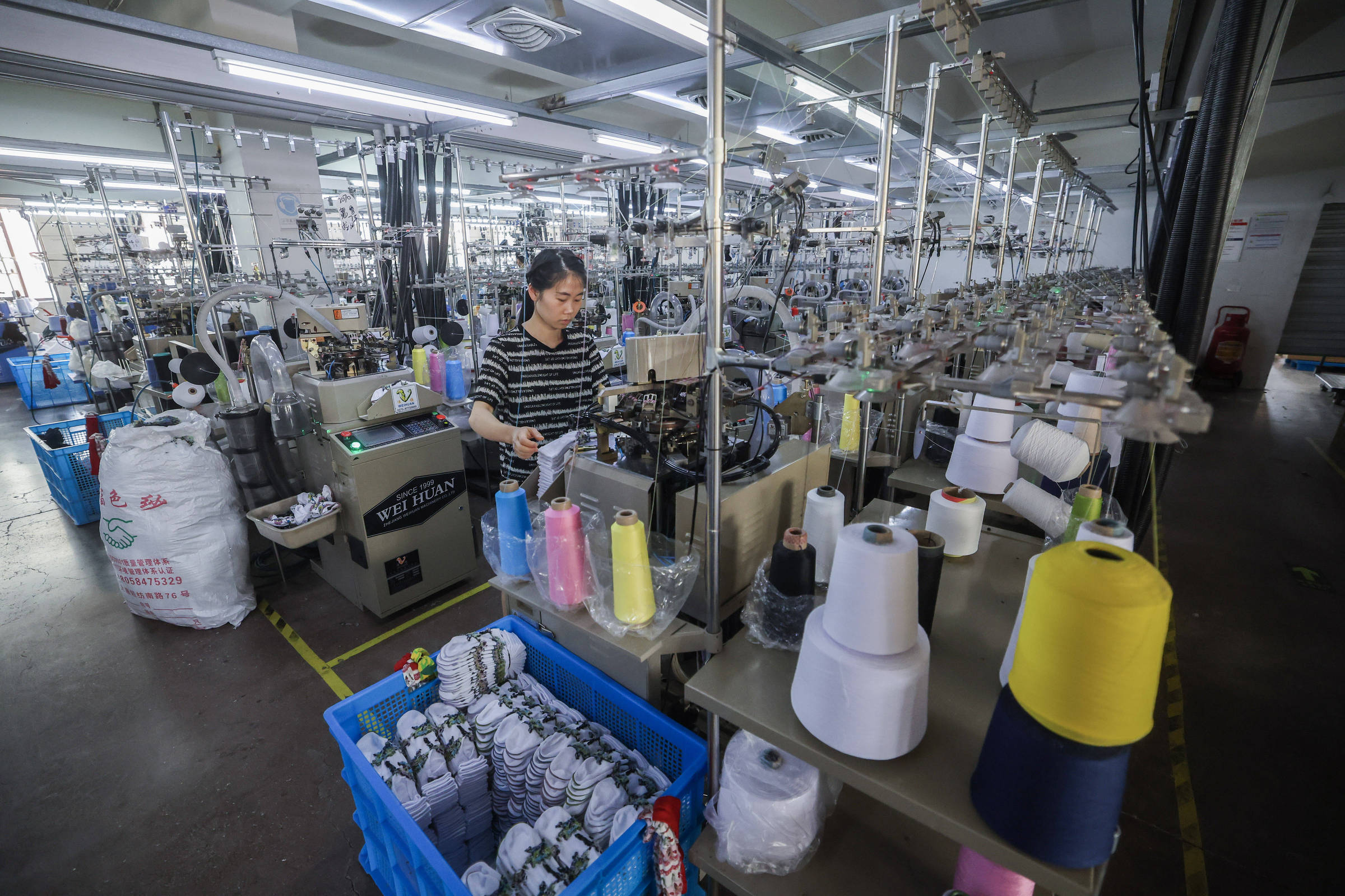 A imagem mostra o interior de uma fábrica de tecidos, com várias máquinas de costura dispostas em fileiras. Uma mulher está operando uma das máquinas, enquanto ao redor há carretéis de fios em diversas cores, organizados em prateleiras e cestos. O ambiente é bem iluminado e parece ser um espaço de trabalho ativo.