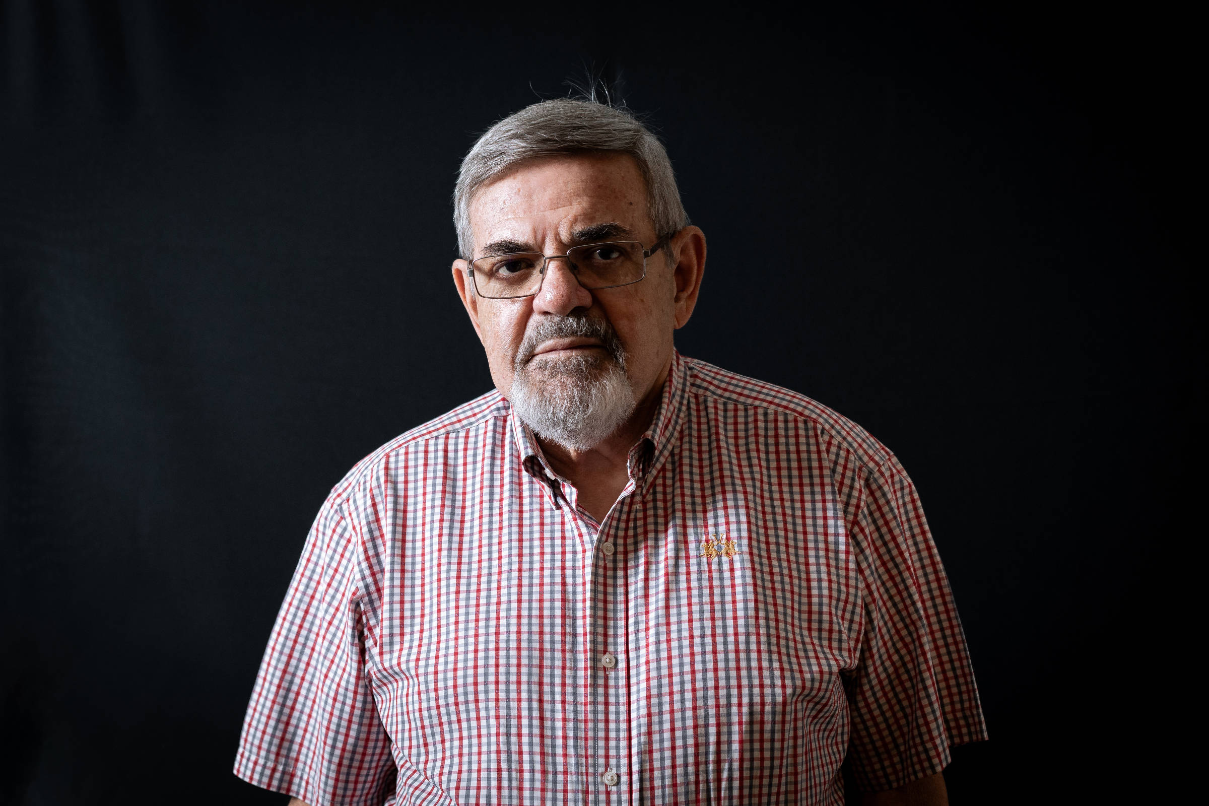 Um homem idoso com cabelo grisalho e barba, usando óculos. Ele está vestido com uma camisa de quadrados vermelhos e brancos. O fundo da imagem é escuro, destacando sua figura. O homem tem uma expressão séria e está olhando diretamente para a câmera.