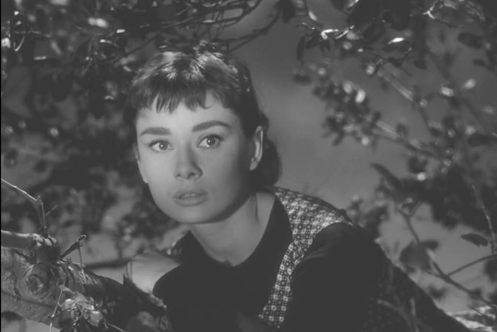 A atriz Audrey Hepburn em um filme preto e branco, vestindo preto e com os cabelos curtos