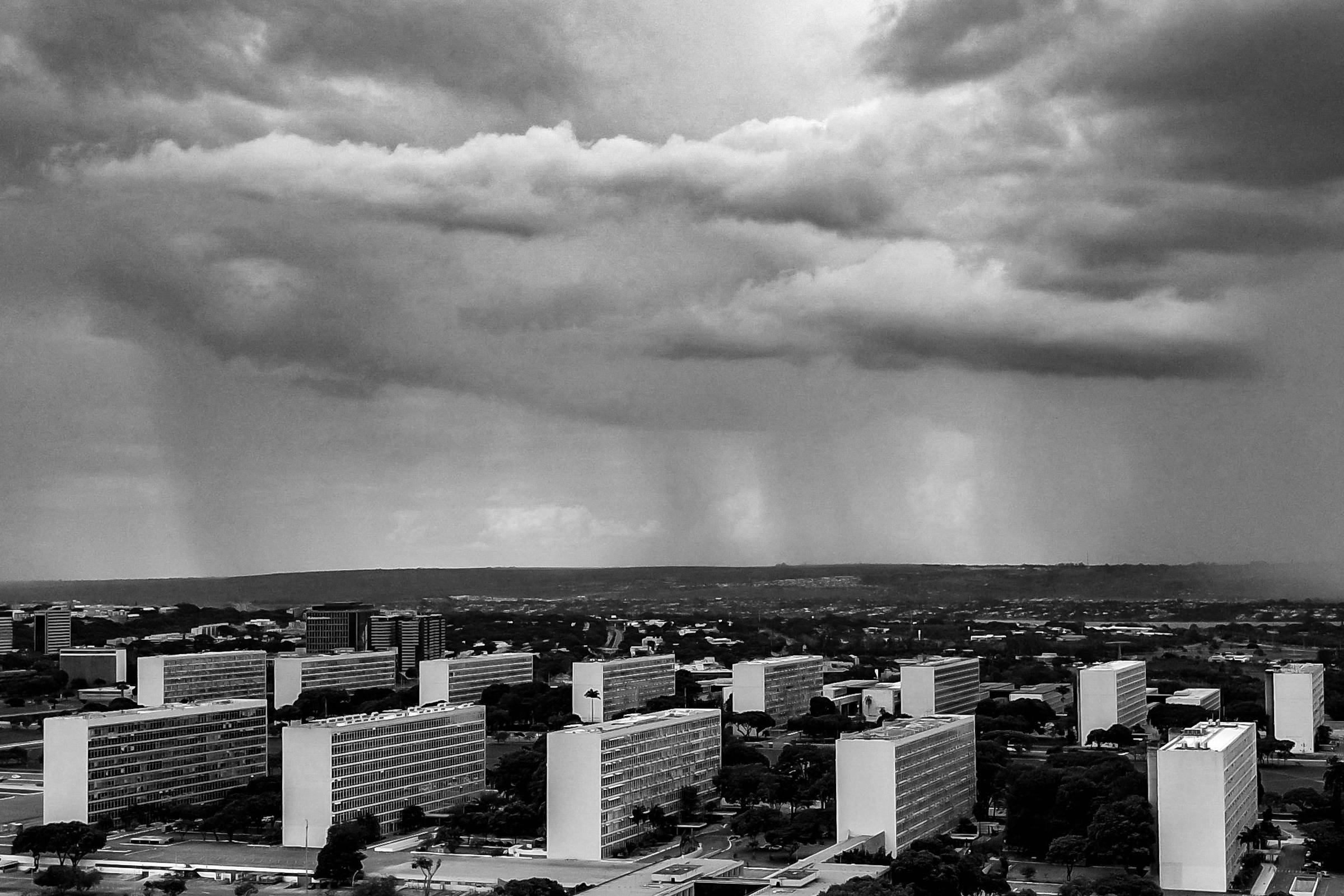 A imagem mostra um céu escuro e nublado, com nuvens pesadas e chuvas caindo em algumas áreas. Abaixo, há uma vista de um conjunto de edifícios baixos e brancos, cercados por áreas verdes. O horizonte é visível ao fundo, onde a chuva parece ser mais intensa.