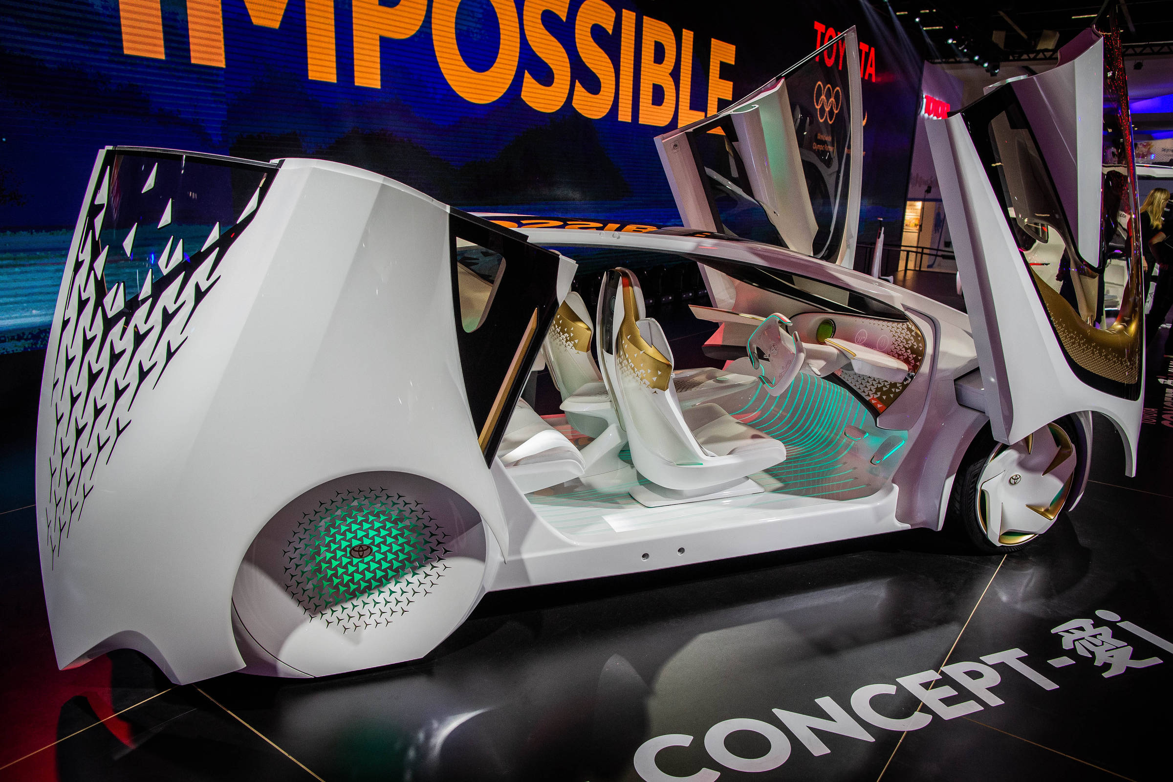 A imagem mostra um carro conceito futurista com um design inovador. O veículo possui portas que se abrem para cima e um interior com assentos brancos e detalhes em cores vibrantes. O exterior é predominantemente branco, com padrões geométricos e elementos de iluminação. Ao fundo, há um painel com a palavra