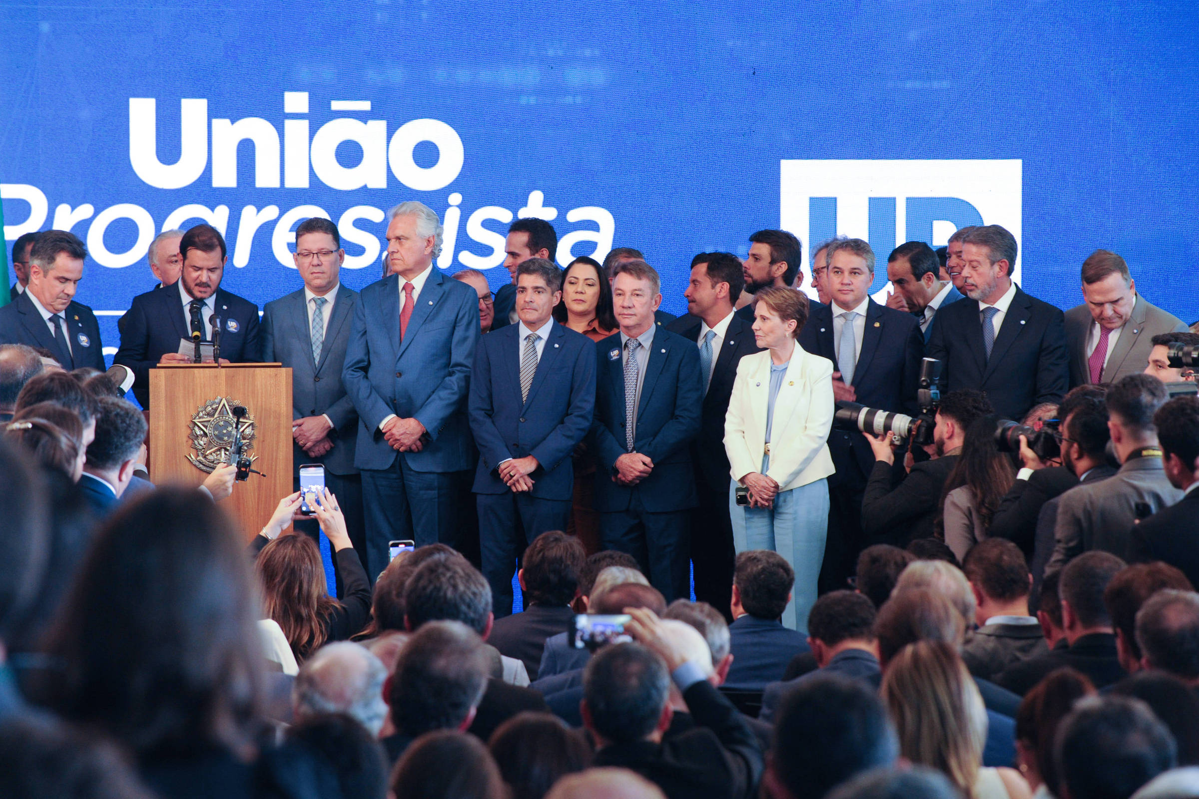 A imagem mostra um evento político com várias pessoas em pé em um palco. Um homem está falando ao microfone em frente a um púlpito, enquanto outros membros do partido estão ao seu lado. Ao fundo, há uma grande tela com o texto