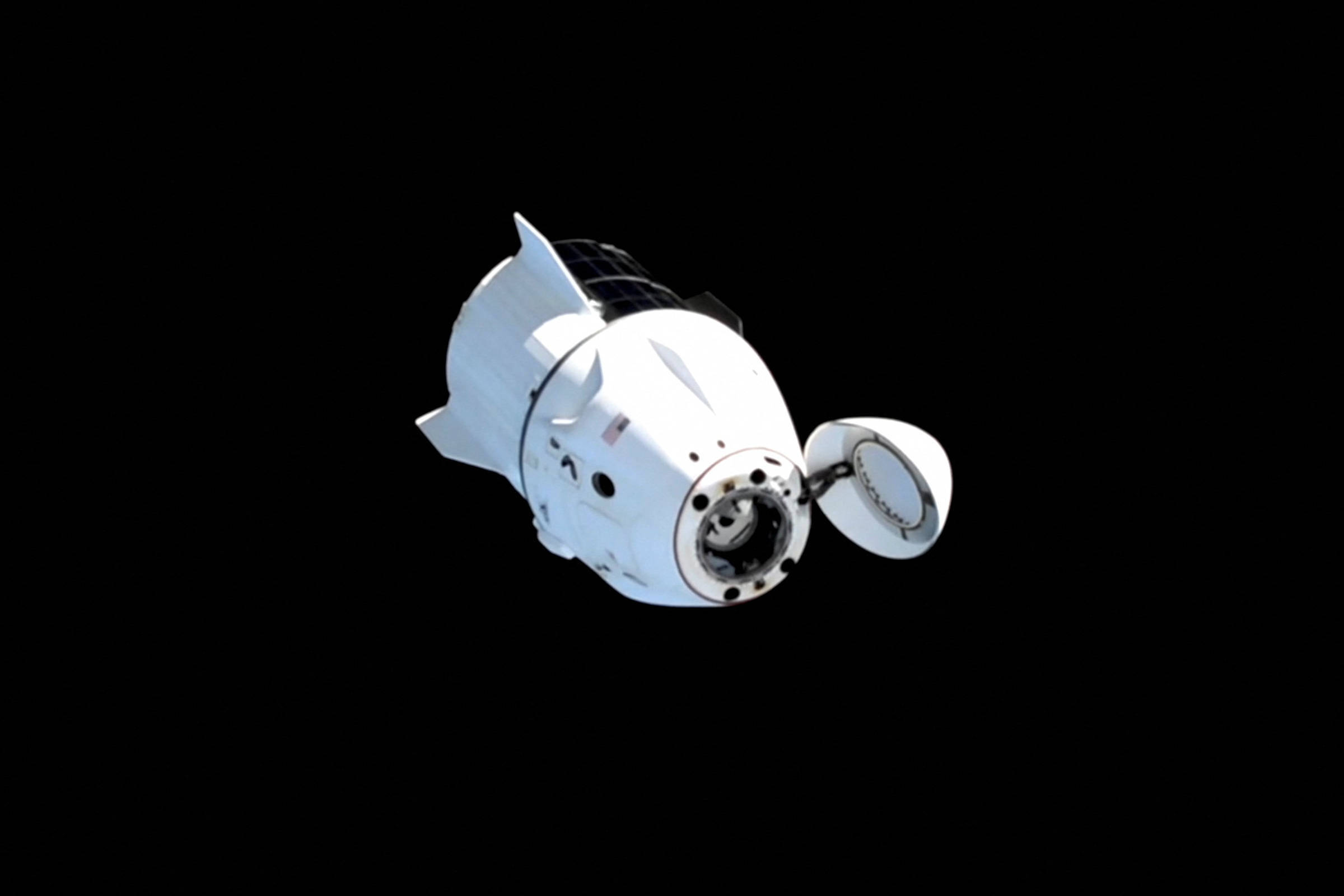 A imagem mostra uma nave espacial branca flutuando no espaço, com um fundo preto. A nave tem uma forma arredondada e apresenta uma abertura na parte frontal, possivelmente uma porta ou escotilha. O logotipo da NASA está visível no canto superior direito.