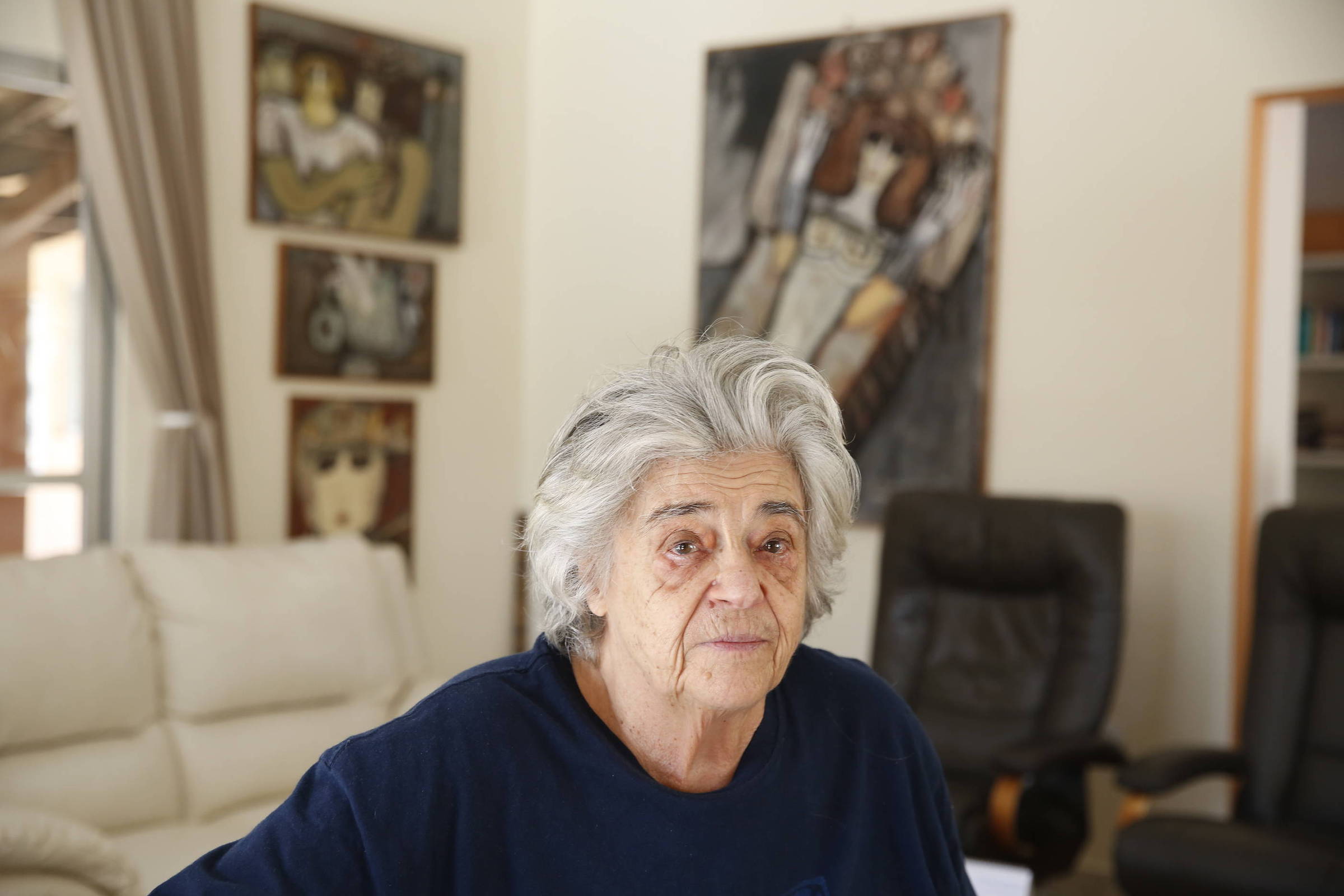 A imagem mostra uma mulher idosa com cabelo grisalho e uma expressão serena, sentada em um ambiente interno. Ao fundo, há várias obras de arte nas paredes, incluindo retratos e uma pintura de uma figura humana. O ambiente é bem iluminado, com um sofá claro e cadeiras escuras visíveis.