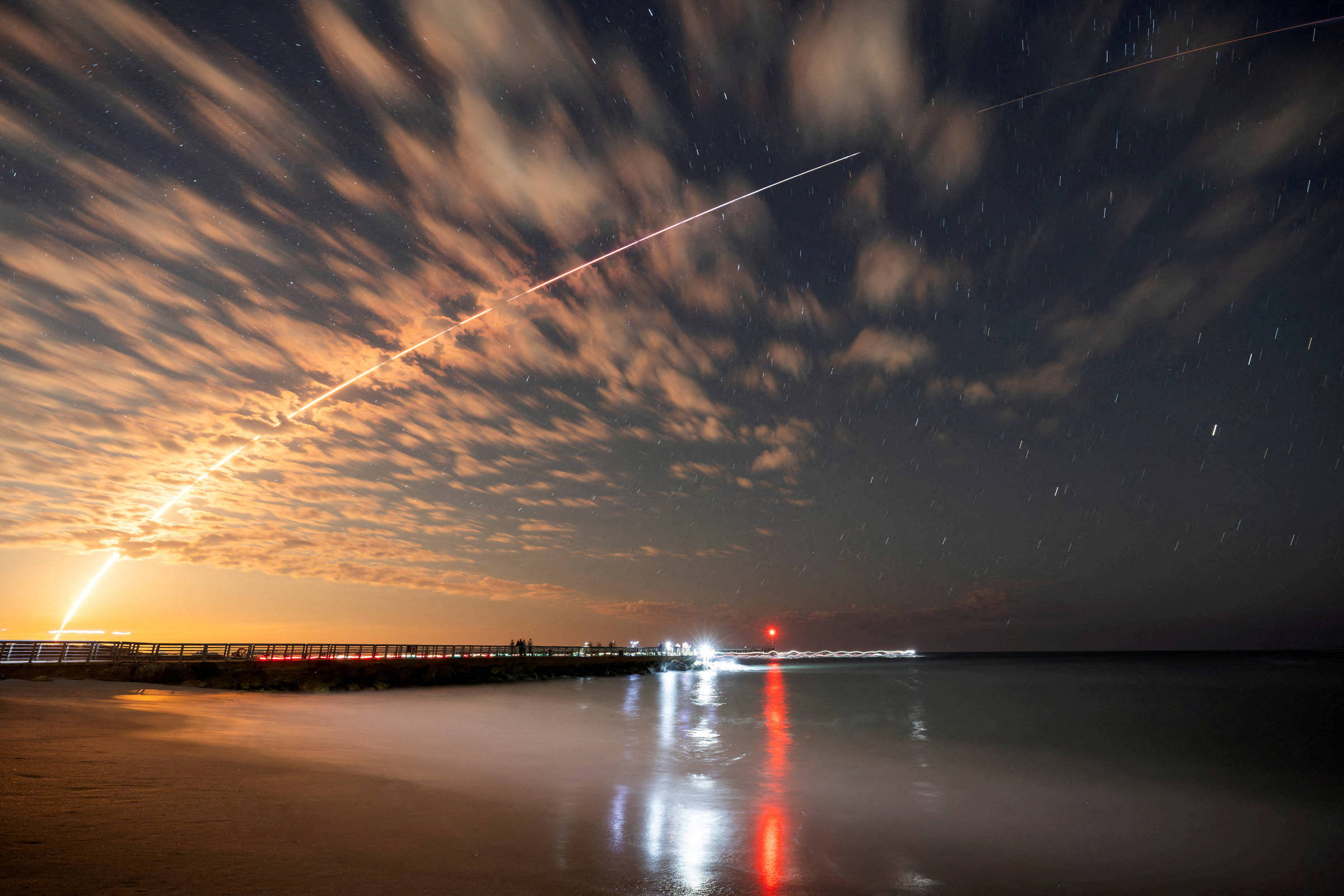 A imagem mostra um céu noturno com nuvens e uma longa trilha de luz que parece ser um foguete ou um projétil sendo lançado. Abaixo, há uma costa com um píer e reflexos de luz na água. À direita, há uma luz vermelha que pode ser um farol ou uma estrutura iluminada. O ambiente é tranquilo, com a água do mar refletindo as luzes.