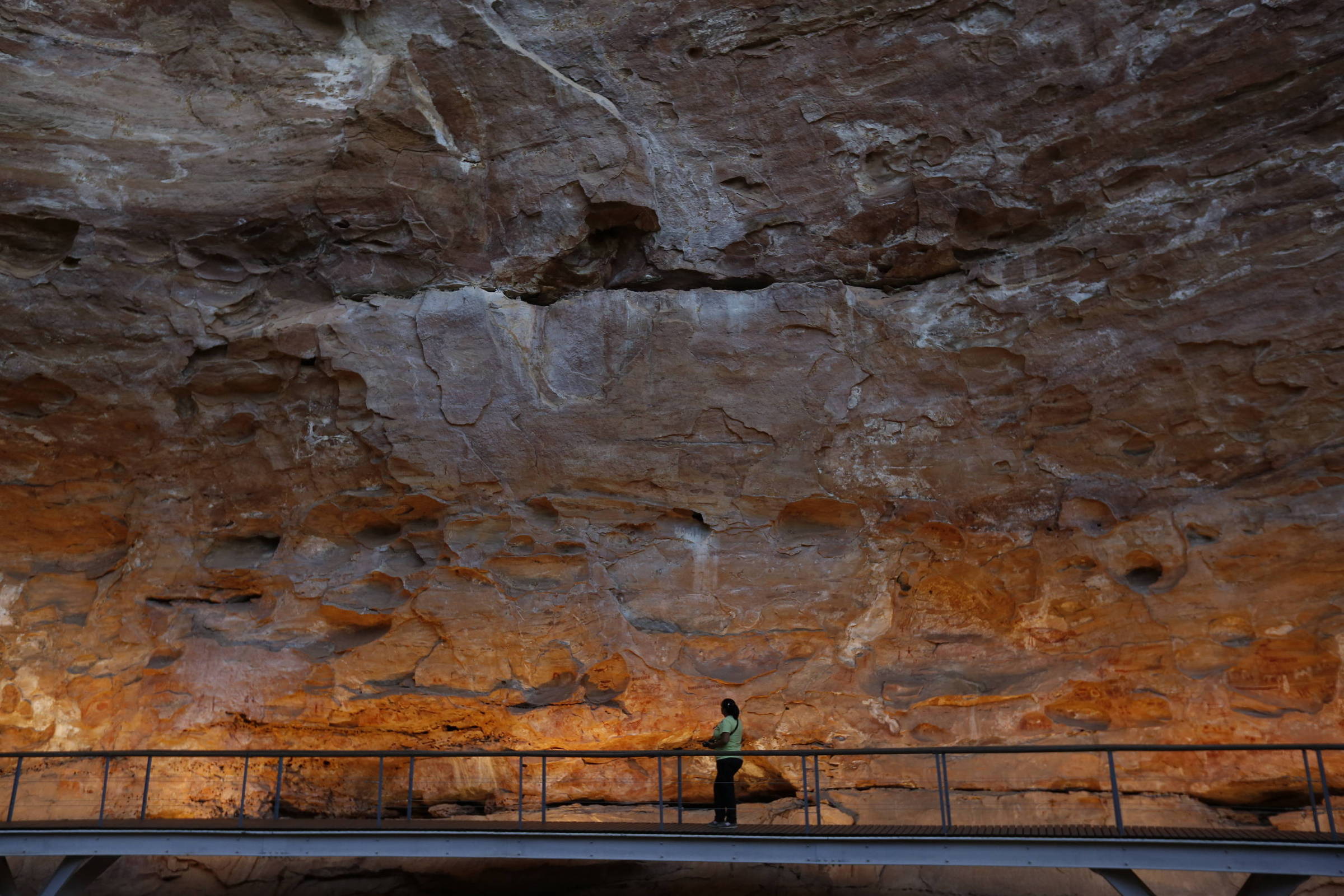 A imagem mostra uma caverna com uma grande parede de rocha exposta. Uma passarela de metal está visível na parte inferior, onde uma pessoa está em pé, observando a formação rochosa. A rocha apresenta várias tonalidades de marrom e laranja, e a iluminação destaca as texturas e as camadas da rocha.