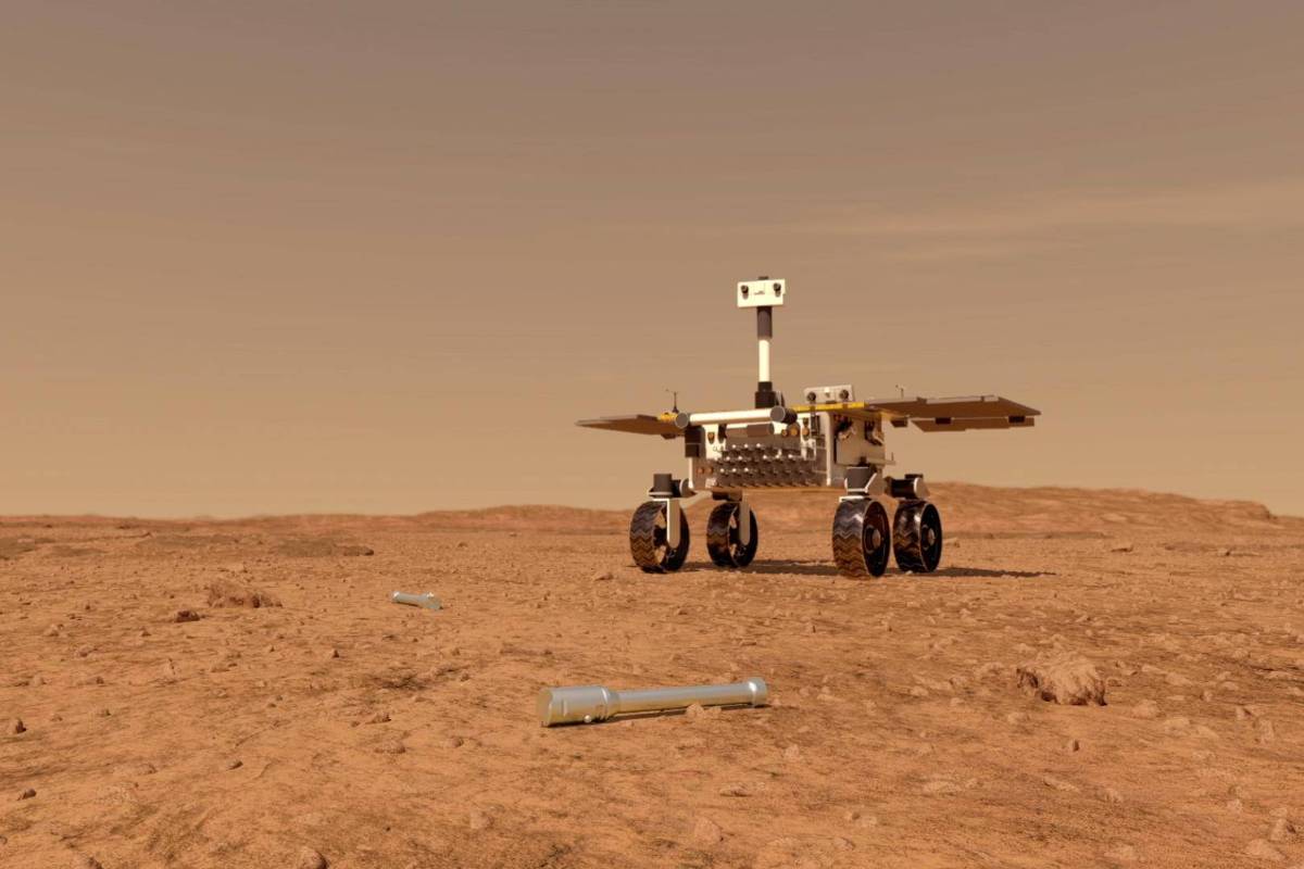 A imagem mostra um rover em uma superfície marciana árida e desértica. O rover possui rodas grandes e uma estrutura com painéis solares. Ao fundo, há uma paisagem de terreno irregular e rochoso, com um céu claro e sem nuvens. No primeiro plano, há alguns objetos metálicos espalhados pelo chão.