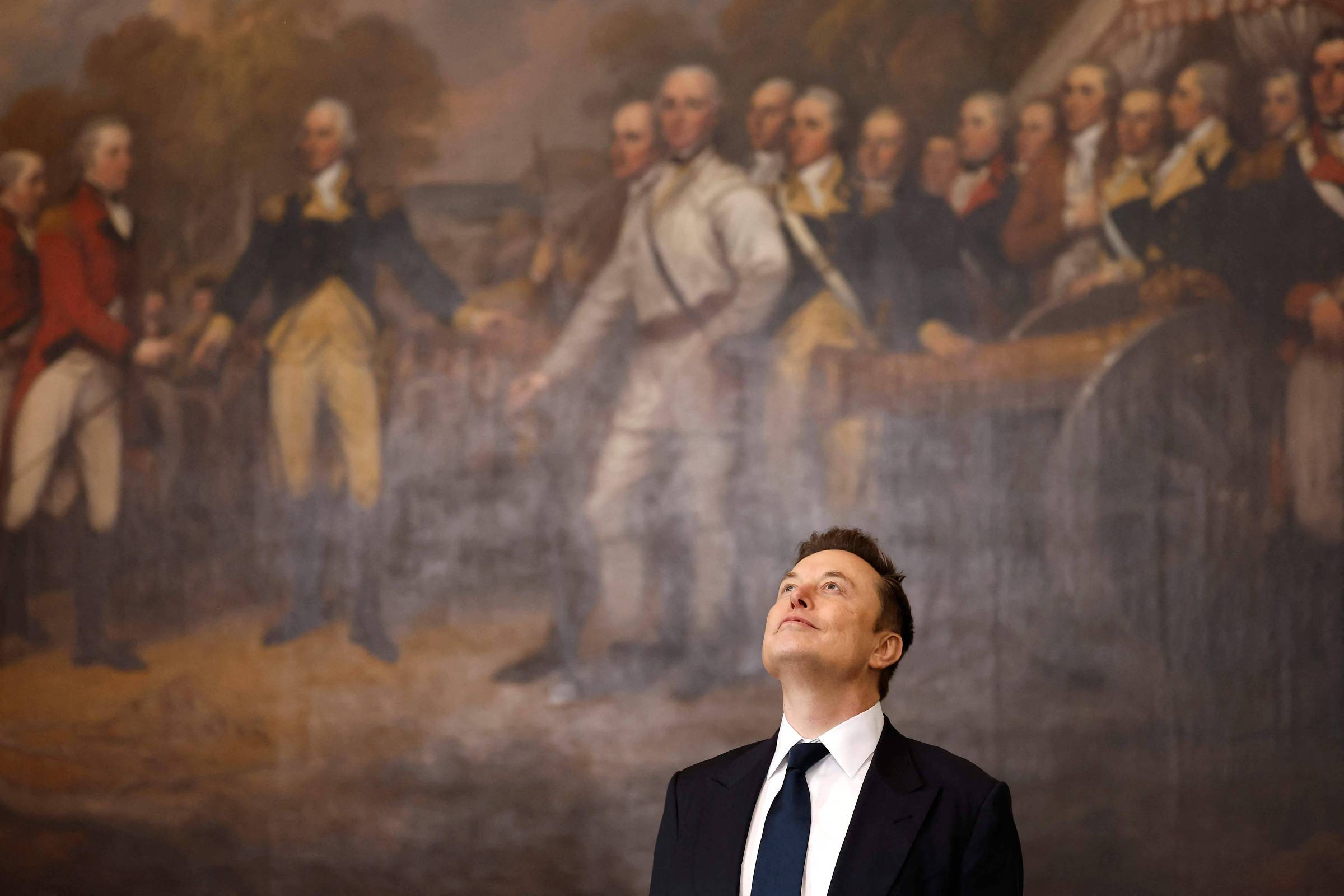 Um homem com terno escuro e gravata branca está olhando para cima, em frente a um grande mural que retrata uma cena histórica com várias figuras em trajes do século XVIII. O mural mostra homens em uniformes militares e civis, em um ambiente que parece ser uma reunião ou um evento significativo.