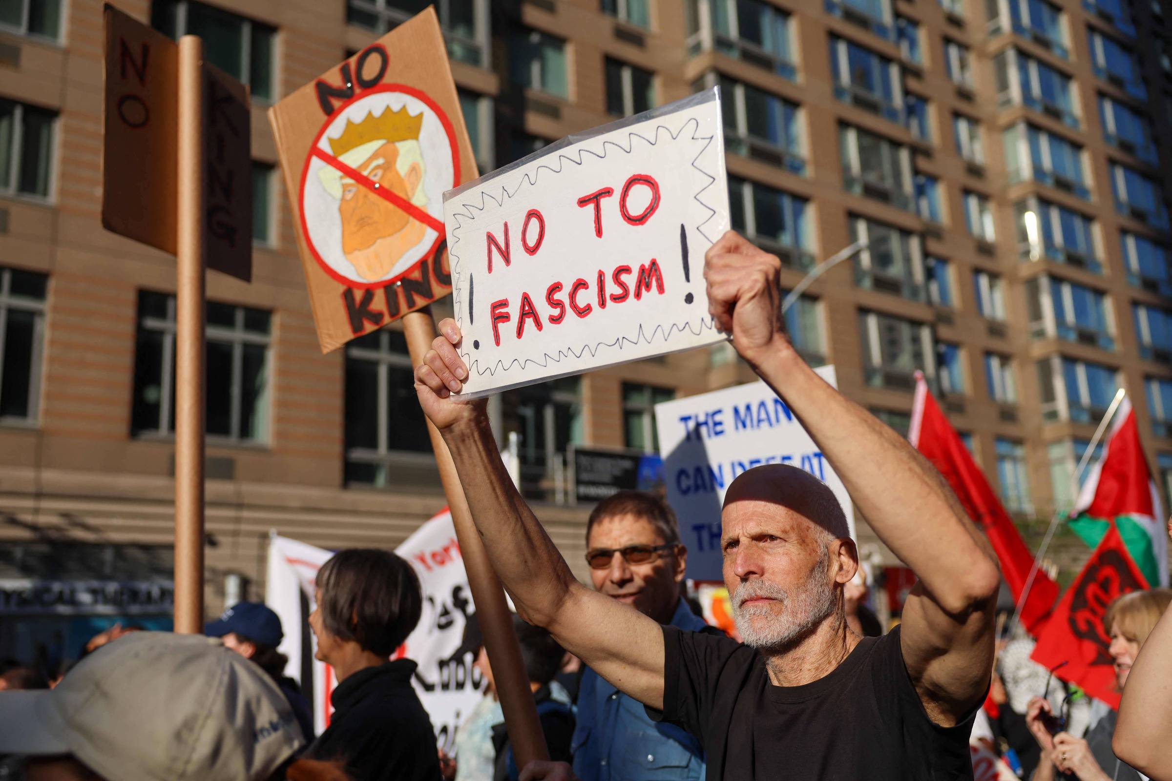 A imagem mostra um grupo de pessoas participando de um protesto. Um homem de cabelo grisalho e barba, vestindo uma camiseta preta, segura um cartaz que diz