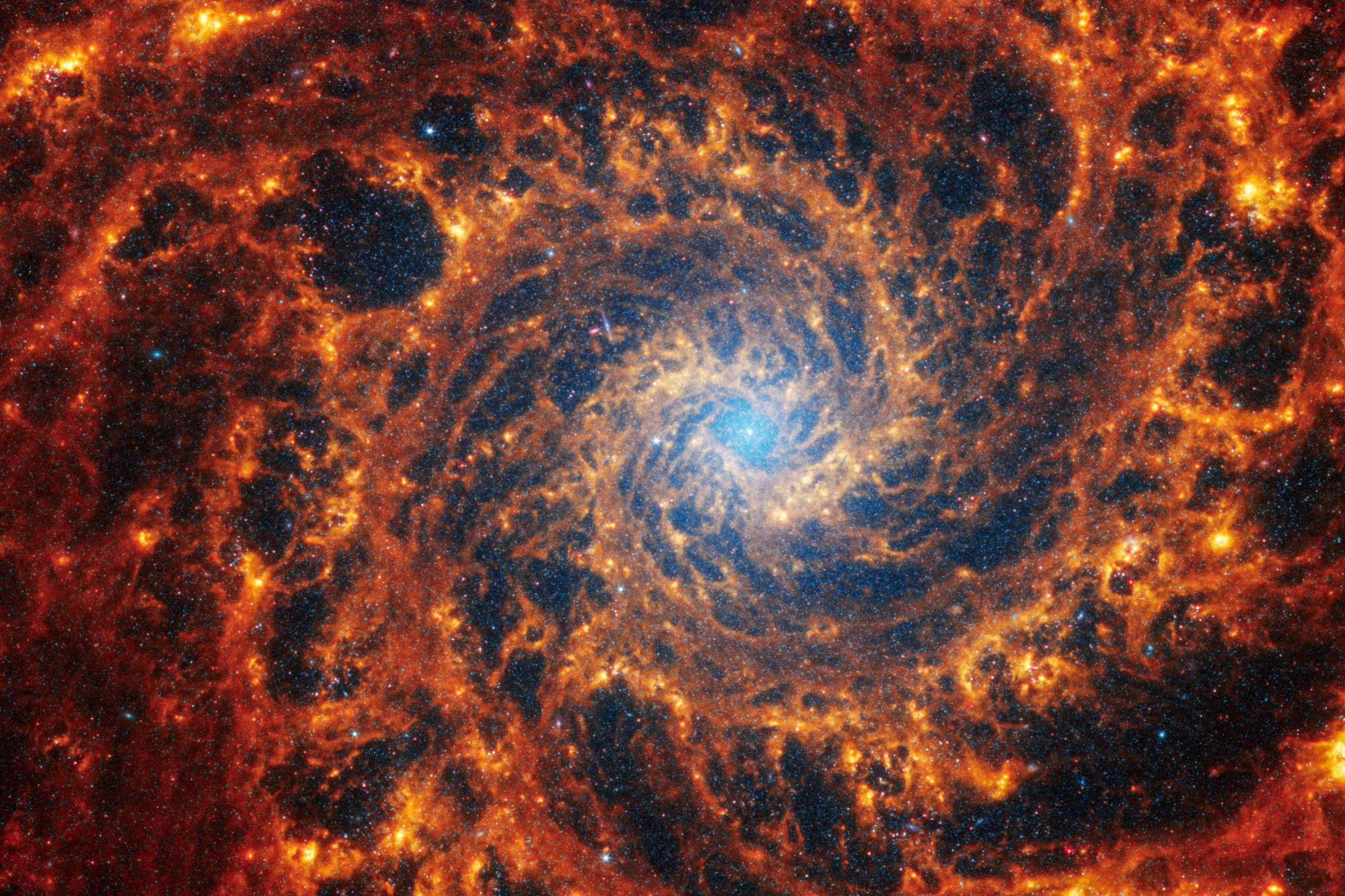 A imagem mostra uma galáxia espiral com um núcleo brilhante em azul, cercado por braços espirais que se estendem em tons de laranja e vermelho.