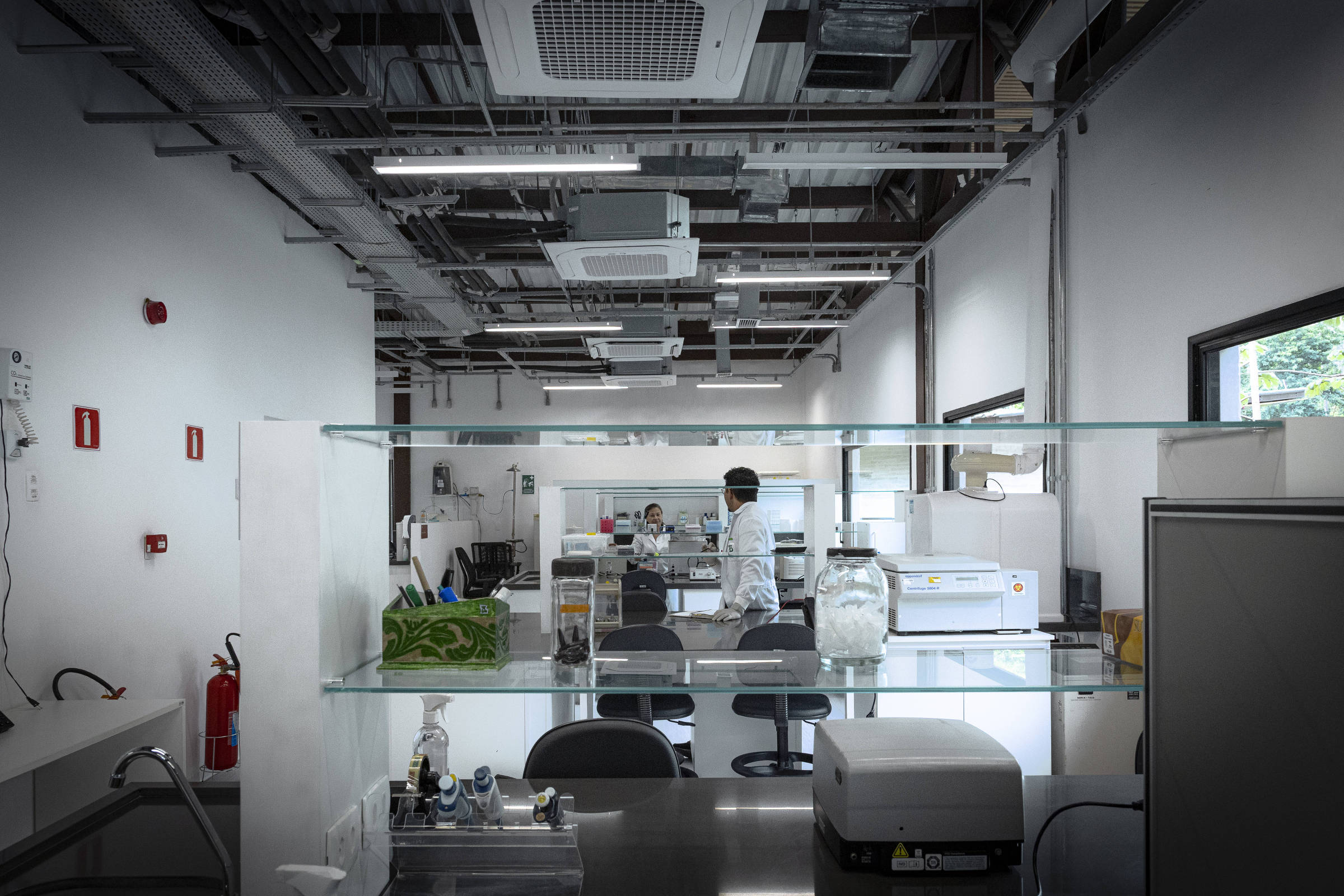 A imagem mostra o interior de um laboratório moderno, com paredes brancas e um teto exposto. Há mesas de trabalho com equipamentos científicos, como geladeiras e instrumentos de laboratório. Uma pessoa vestindo um jaleco está em pé, aparentemente realizando alguma atividade. O ambiente é bem iluminado e organizado, com prateleiras de vidro e uma área de trabalho limpa.