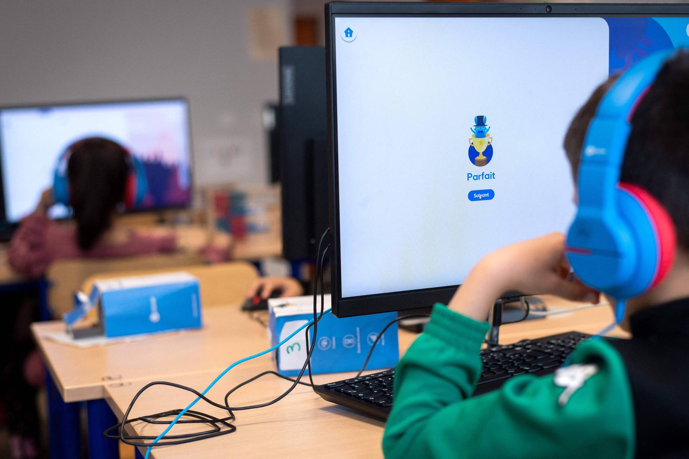 A imagem mostra uma sala de aula de informática com crianças usando computadores. Um menino está sentado em frente a um monitor, usando fones de ouvido azuis. Na tela do computador, aparece o logotipo de um aplicativo chamado