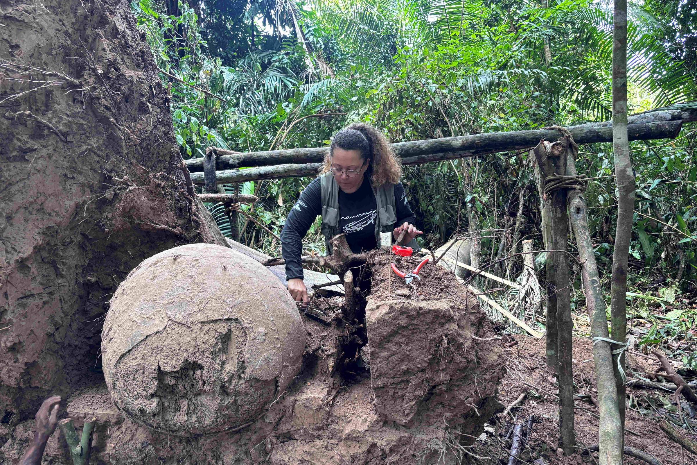 Uma pessoa está trabalhando em uma área de floresta densa, próxima a uma grande esfera de pedra parcialmente enterrada. A esfera é de cor clara e apresenta marcas na superfície. A pessoa, com cabelo cacheado e preso, está usando uma roupa escura e parece estar utilizando ferramentas para escavar ao redor da esfera. Ao fundo, há vegetação densa e uma estrutura de madeira que parece ser um suporte improvisado.