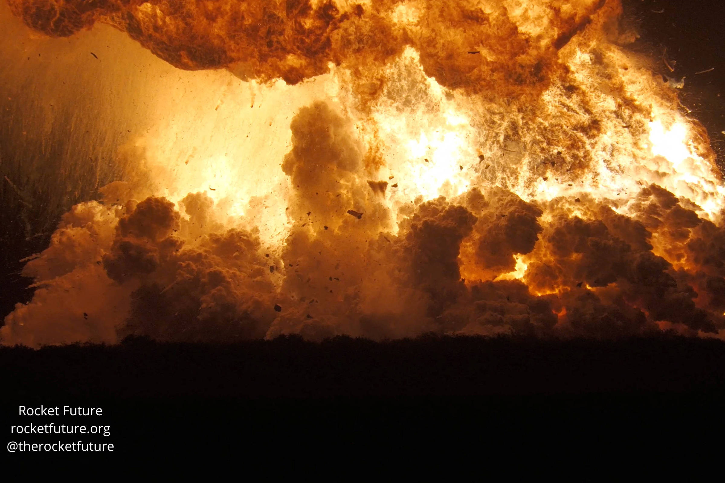 A imagem mostra uma explosão intensa com chamas e fumaça. O fogo é predominante, criando uma nuvem de fumaça laranja e amarela, enquanto fragmentos são lançados para o ar. A parte inferior da imagem é escura, contrastando com a luminosidade da explosão. Há texto no canto inferior esquerdo, que inclui o nome