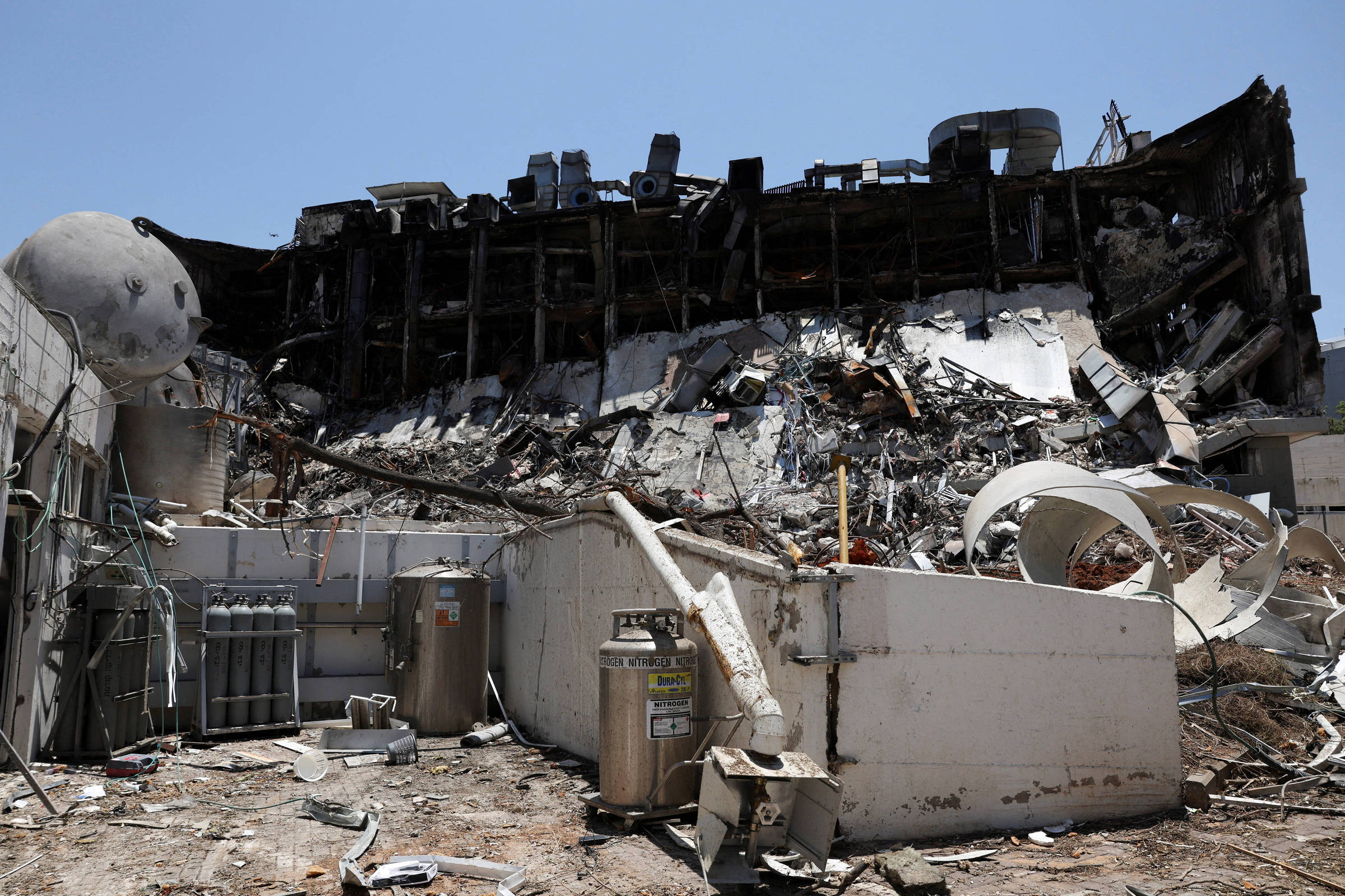 A imagem mostra a estrutura de um edifício em colapso, com a parte superior desmoronada e destroços espalhados pelo chão. Há equipamentos e materiais de construção visíveis entre os escombros, e o céu está claro ao fundo.