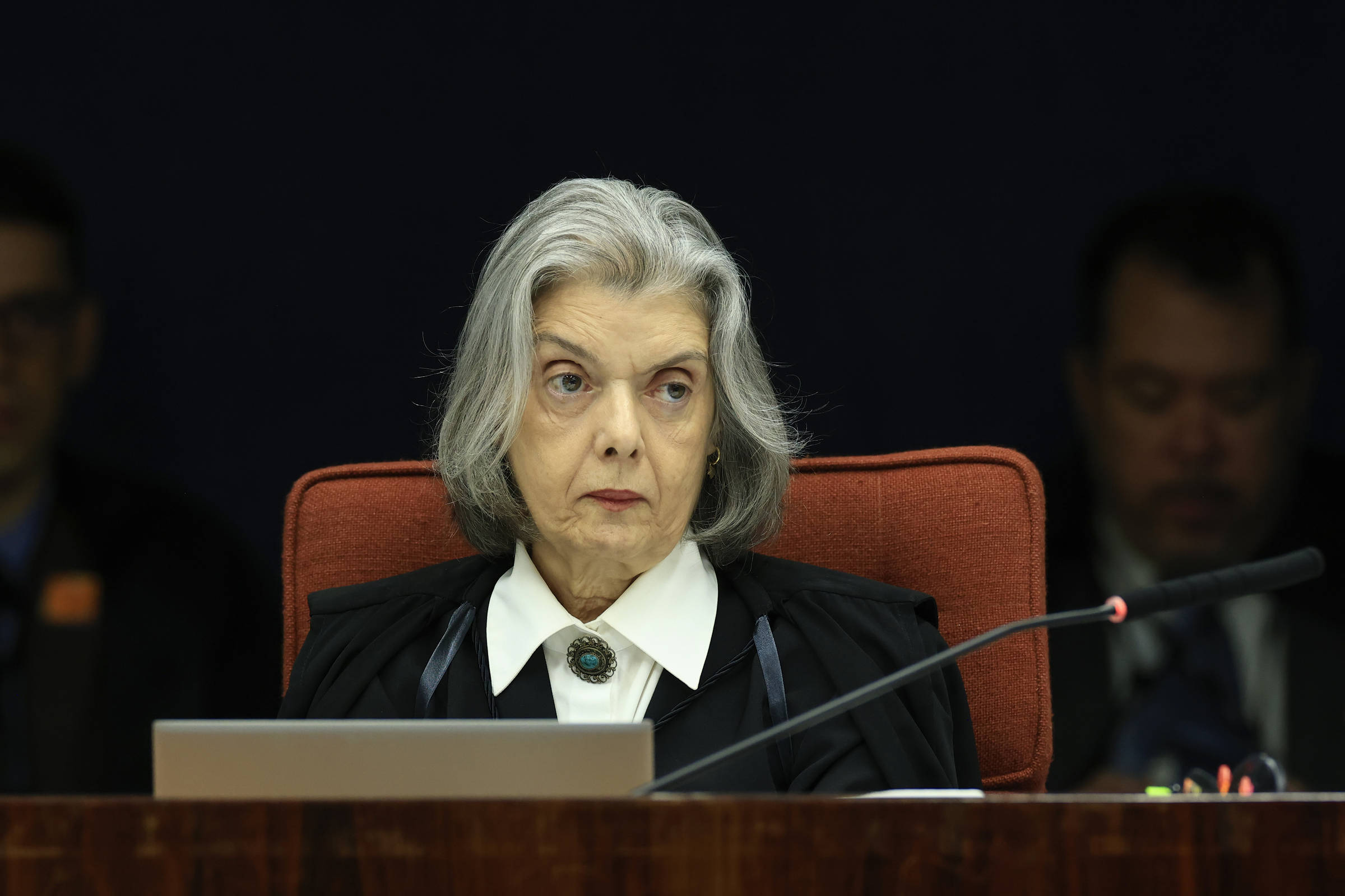 Uma juíza com cabelo grisalho e liso, vestindo uma toga preta e uma blusa branca, está sentada em uma mesa de tribunal. Ela parece concentrada e séria, olhando para frente. Ao fundo, há pessoas indistintas, e a mesa possui um microfone e outros objetos.