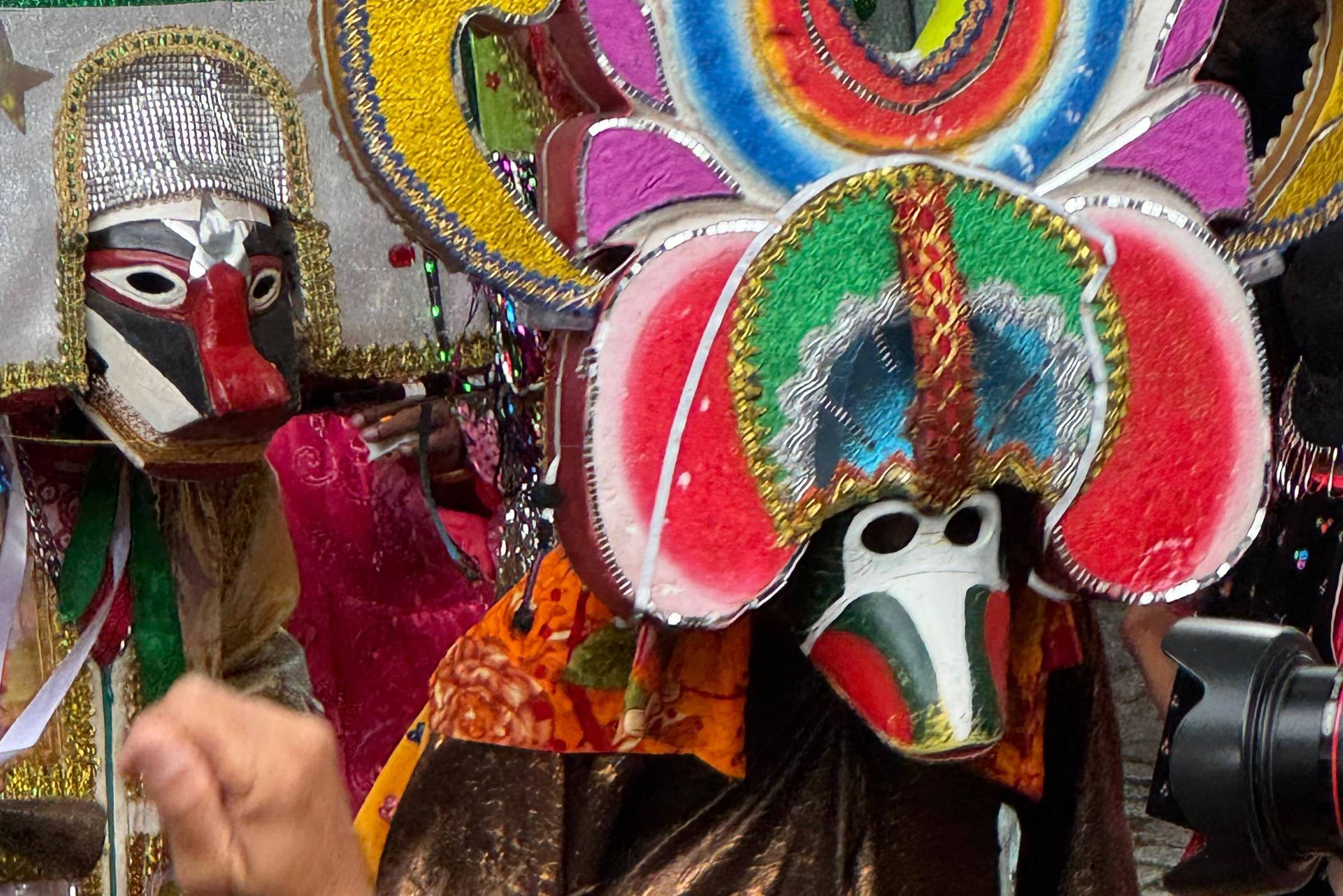 A imagem mostra um grupo de pessoas participando de uma festa, onde um dos indivíduos está usando uma máscara elaborada e colorida, adornada com penas e brilhos. Ao fundo, é possível ver outras pessoas e elementos decorativos típicos de festividades. Um cartaz com uma imagem religiosa é visível ao fundo, complementando o cenário festivo.