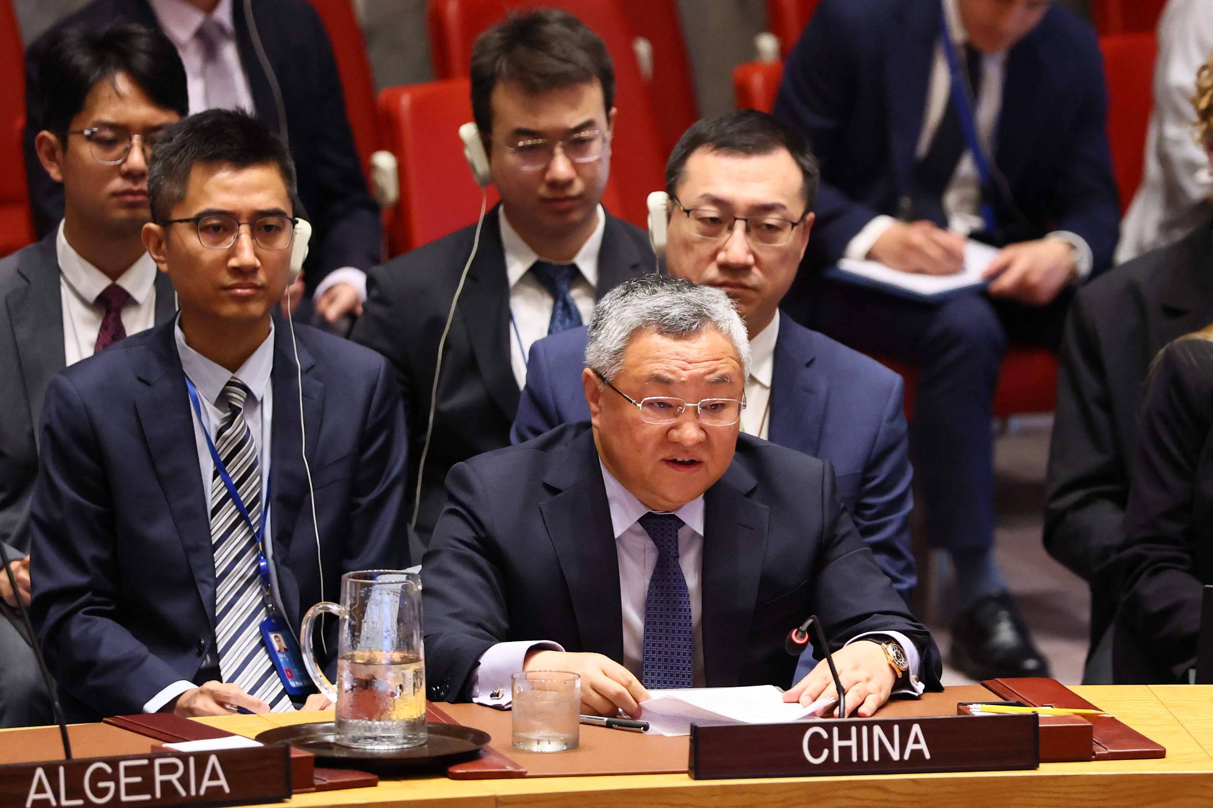 A imagem mostra uma reunião do Conselho de Segurança da ONU, onde um homem de terno escuro e óculos está falando ao microfone, representando a China. Atrás dele, há vários outros homens em trajes formais, alguns usando fones de ouvido. Na mesa, há placas com os nomes dos países, incluindo