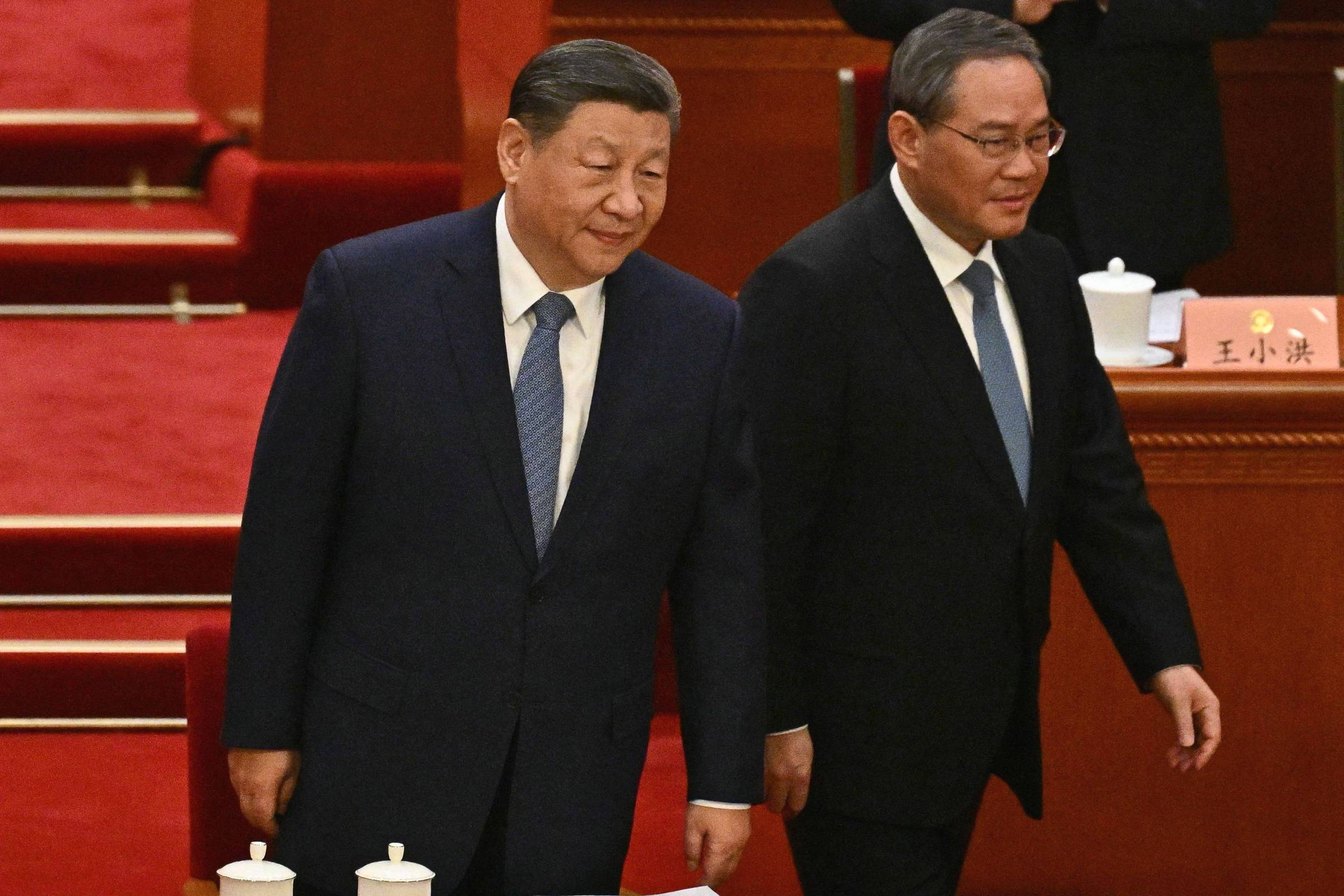 Xi Jinping e Li Qiang, dois homens em trajes formais, caminham juntos em um ambiente de evento oficial. Xi, o homem à esquerda, tem cabelo curto e está usando um terno escuro com uma gravata azul. Li Qiang, o homem à direita, também está vestido formalmente, com um terno escuro e gravata preta. O fundo é decorado com mesas e copos brancos, e há um tapete vermelho no chão.