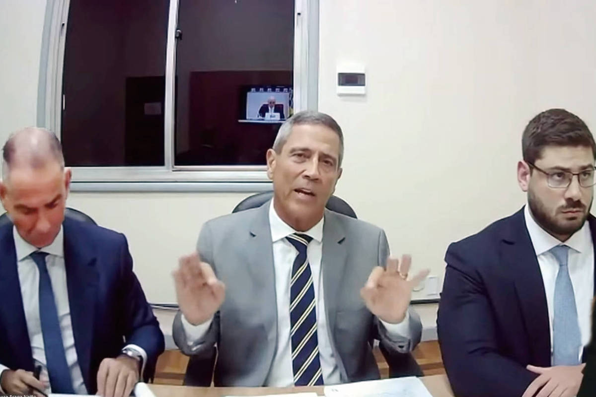 A imagem mostra três homens sentados em uma mesa em um ambiente de escritório. O homem do meio, vestido com um terno cinza e gravata listrada, está gesticulando com as mãos enquanto fala. À sua esquerda, um homem de terno escuro e gravata clara observa atentamente. À direita, um homem com óculos e barba, também de terno escuro, parece concentrado. Ao fundo, uma tela exibe uma imagem de outra reunião ou apresentação.