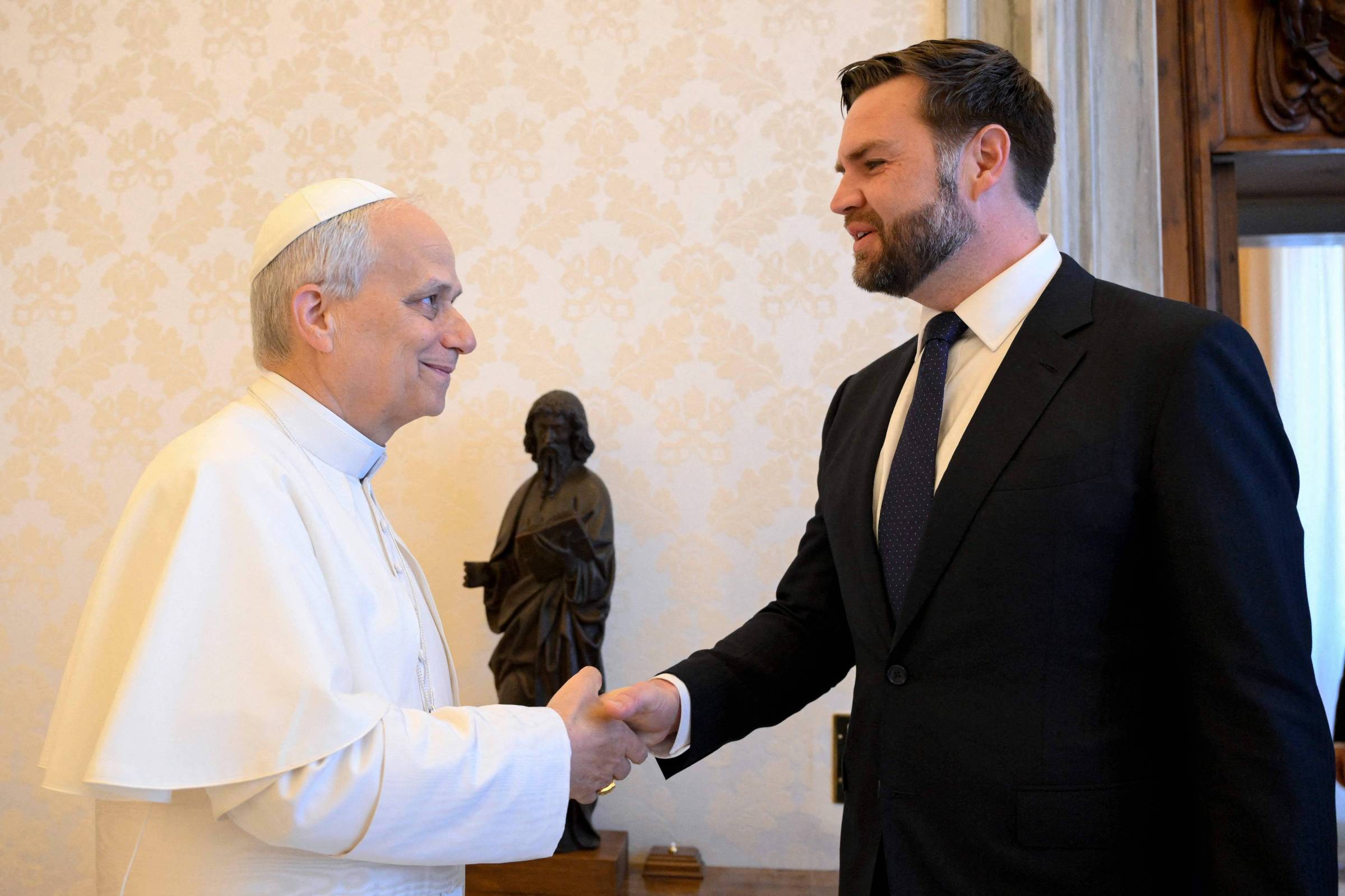 A imagem mostra um encontro entre duas pessoas, uma delas vestida com uma batina branca, representando o Papa, e a outra em um terno escuro. Eles estão se cumprimentando com um aperto de mão. Ao fundo, há uma escultura de uma figura humana em pé. O ambiente é interno, com paredes decoradas e uma atmosfera formal.