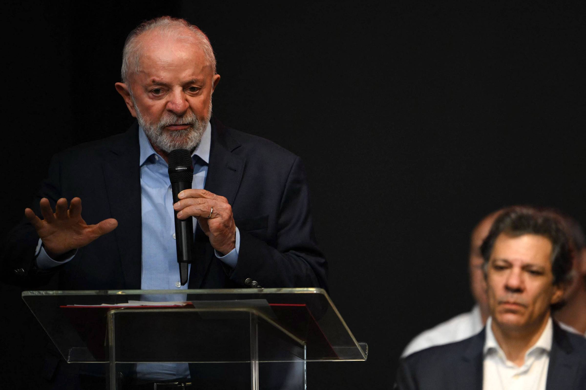 Um homem de cabelo grisalho e barba, vestido com um terno escuro, está em pé atrás de um púlpito de vidro, falando ao microfone. Ele gesticula com a mão direita enquanto se dirige a uma audiência. Ao fundo, um homem com cabelo escuro e camisa clara observa atentamente. O ambiente é escuro, com foco na figura do orador.