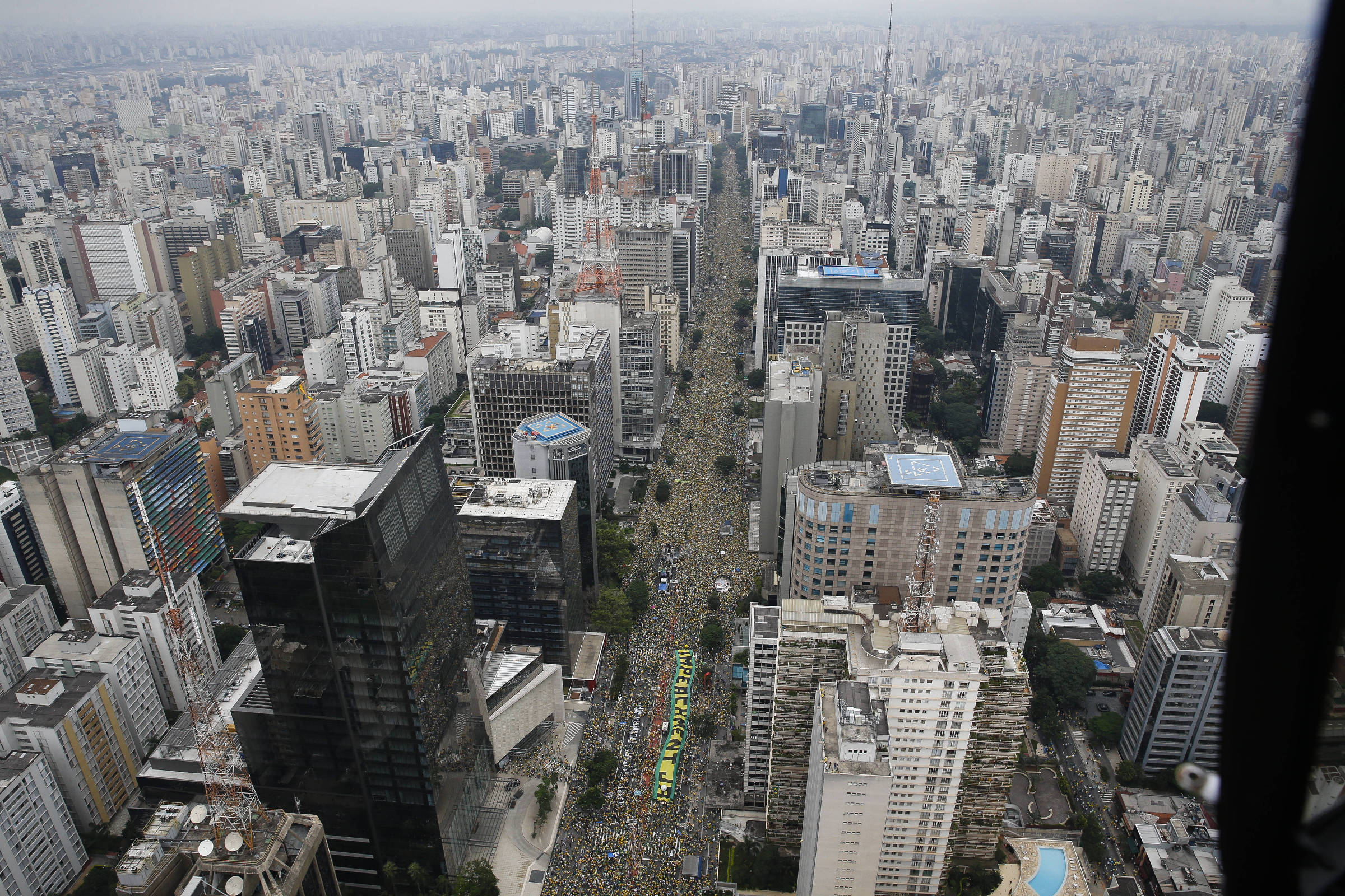A imagem mostra uma vista aérea da cidade de São Paulo, com uma grande quantidade de prédios e arranha-céus. No centro da imagem, uma avenida está repleta de pessoas participando de uma manifestação, com bandeiras e cartazes visíveis. O céu está nublado, e a cidade parece densa, com muitos edifícios de diferentes tamanhos e estilos arquitetônicos.