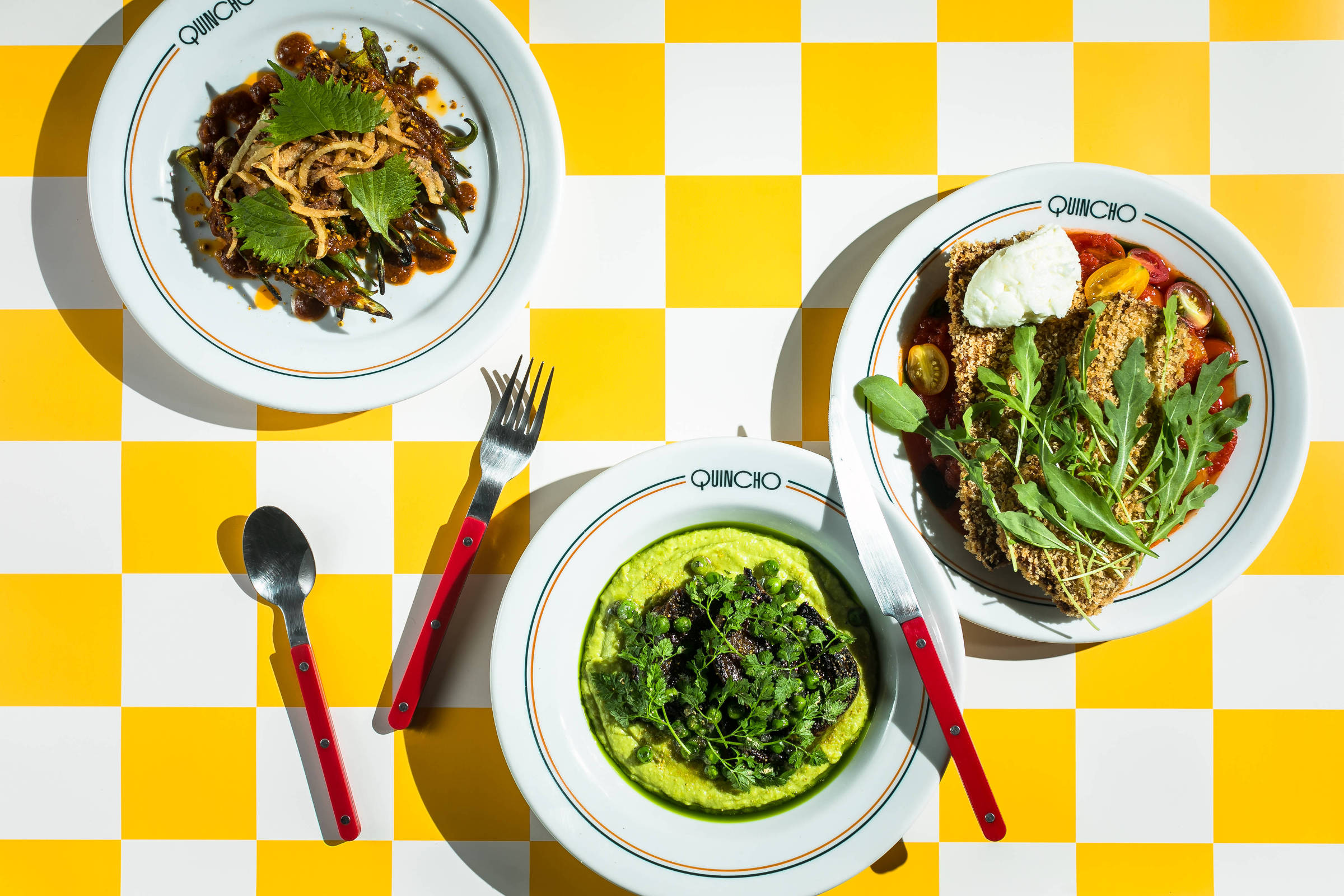 A imagem mostra três pratos de comida dispostos sobre uma mesa com toalha xadrez amarela e branca. O prato à esquerda contém uma mistura de vegetais e ervas. O prato central apresenta um purê verde com folhas escuras por cima. O prato à direita é uma salada com vegetais variados e um ovo cozido. Os talheres, com cabos vermelhos, estão posicionados ao lado dos pratos.