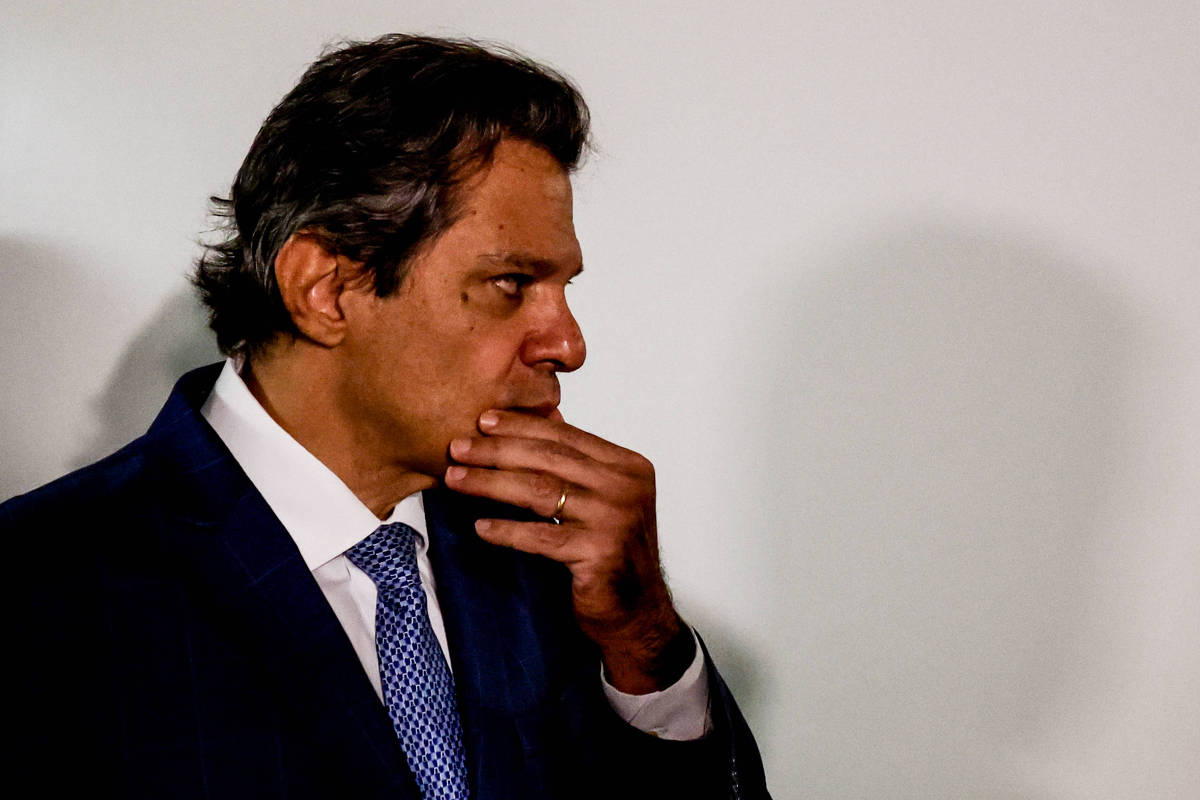 Um homem com cabelo escuro e levemente ondulado está em perfil, olhando para baixo. Ele usa um terno azul escuro com uma camisa branca e uma gravata azul. O homem está com a mão na boca, em uma expressão pensativa. O fundo é claro e neutro, destacando sua figura.